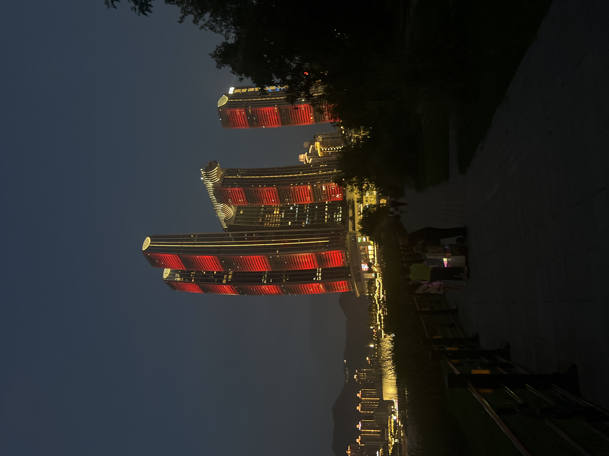 The image depicts a nighttime scene in Jiangbeicheng Subdistrict, China, featuring a striking architectural complex illuminated with red and yellow lights. The foreground shows a paved pathway with a black metal railing on the left side, bordered by dark vegetation. Several silhouetted figures are visible on the pathway, some appearing to be a small group gathered around a stall. In the mid-ground, a waterfront area is lit with numerous yellow lights, suggesting buildings and possibly a promenade.
Dominating the background are several skyscrapers. The most prominent are two tall buildings with their upper sections lit in alternating horizontal bands of bright red and yellow. Another taller, slender building to the right is also illuminated with yellow lights. Further in the distance, a cityscape with more illuminated buildings and faint lights on a hillside can be seen, with dark hills forming the horizon. The sky is a deep, dark blue, characteristic of night. The overall impression is one of a vibrant, modern urban landscape at night.