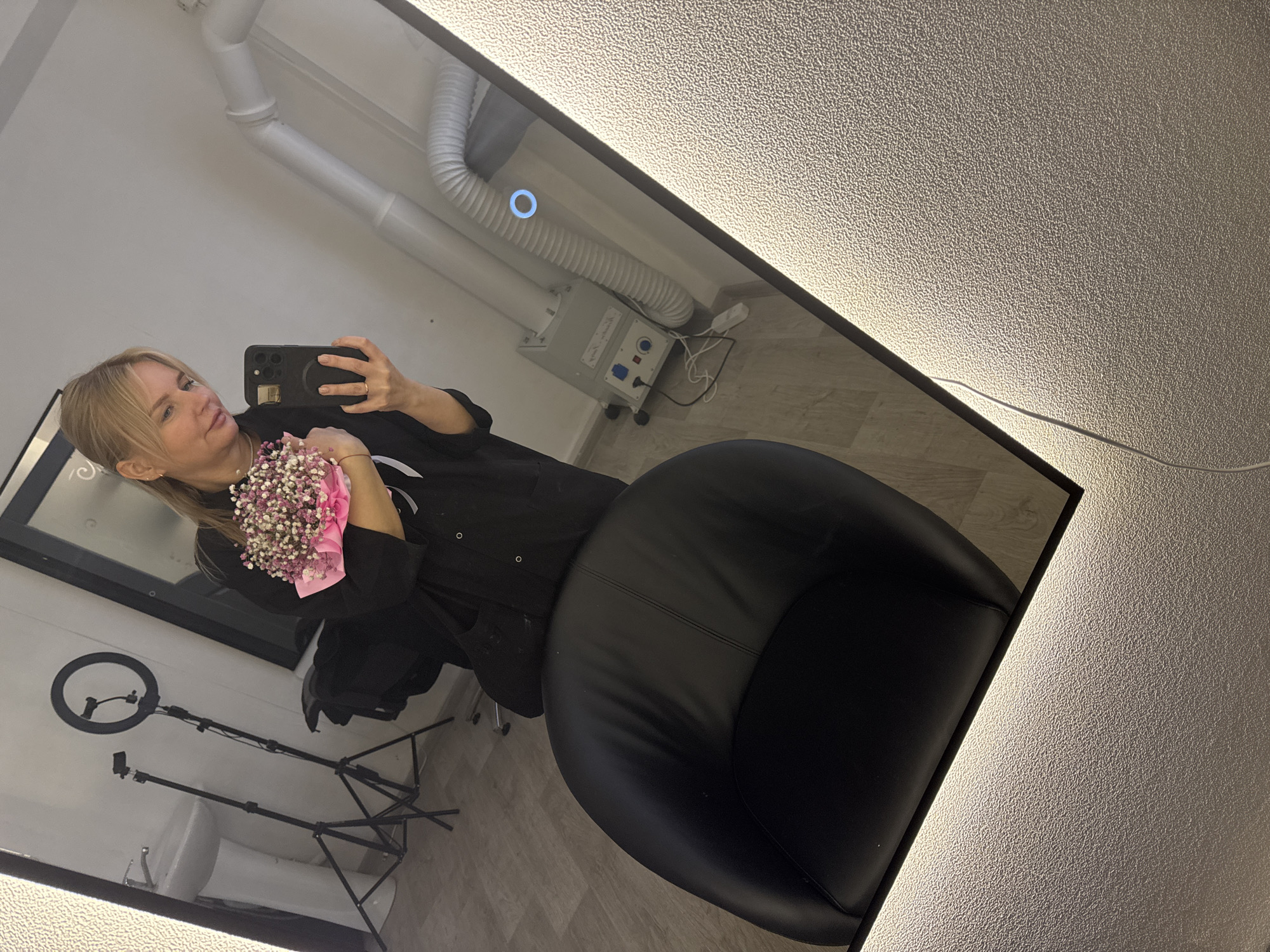 A mirrored selfie is taken by a blonde woman in what appears to be a salon or similar establishment. The reflection shows a woman holding a bouquet of small white and pink flowers wrapped in pink paper. She is dressed in a dark, possibly black garment. The reflection also reveals elements of the room, including a black chair, a ring light on a stand, a piece of medical equipment with hoses, and pipes on the ceiling. The mirror is illuminated along its edges. Due to the lack of details and without geolocation services, I cannot pinpoint to a specific location in Chaikovsky, Russia.