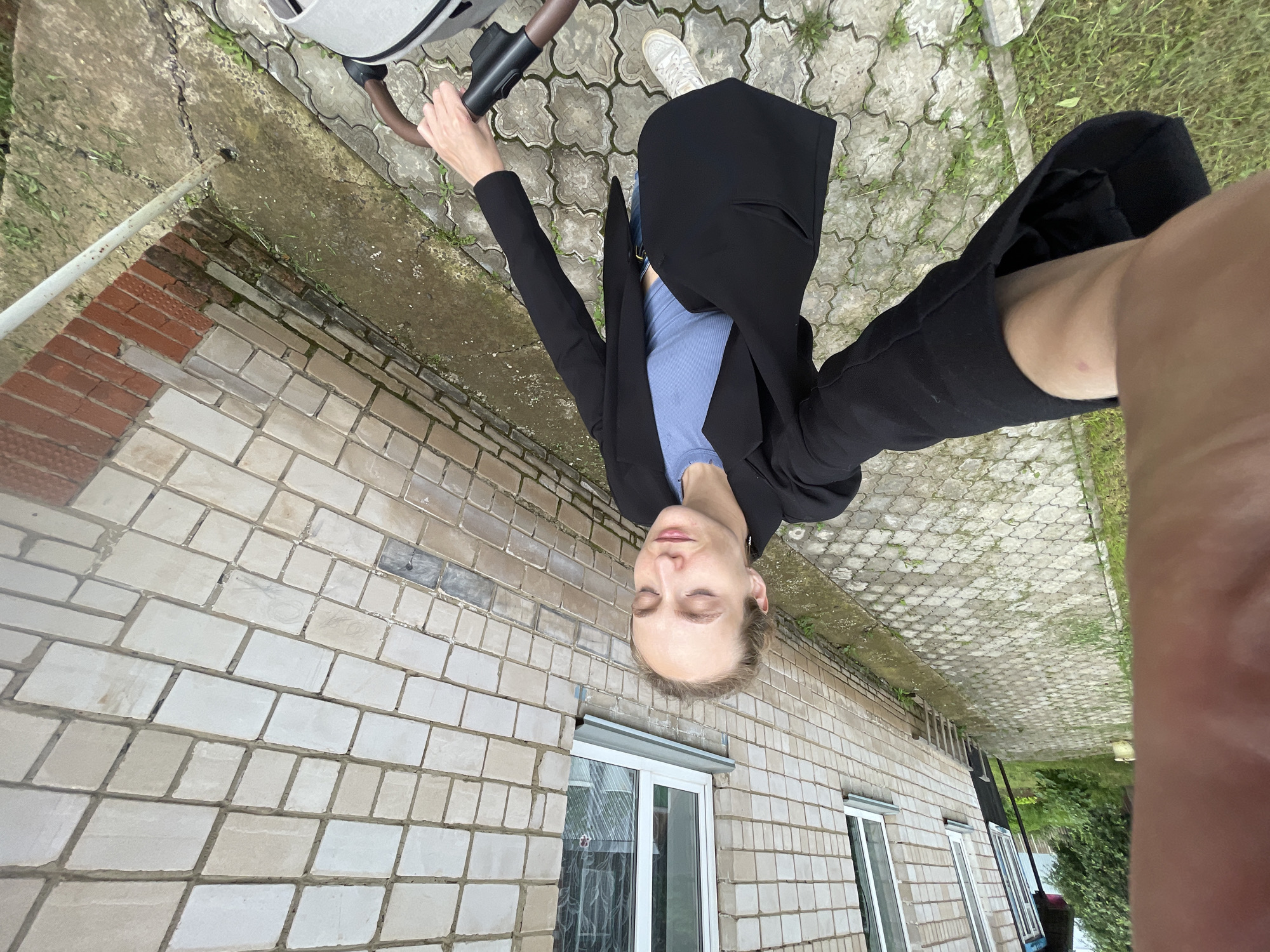 In the city of Sharkan, Russia, a woman is pictured in a selfie. She is standing on a patterned concrete walkway next to a building constructed of light-colored brick. The woman is wearing a black blazer over a blue top and blue jeans, with white sneakers on her feet. Her eyes are closed, and she appears to be holding the handle of a baby stroller with her right hand, which is visible in the lower right portion of the frame. The background shows the brick building with windows and some overgrown green vegetation. The image is taken from a high angle, looking down at the woman.