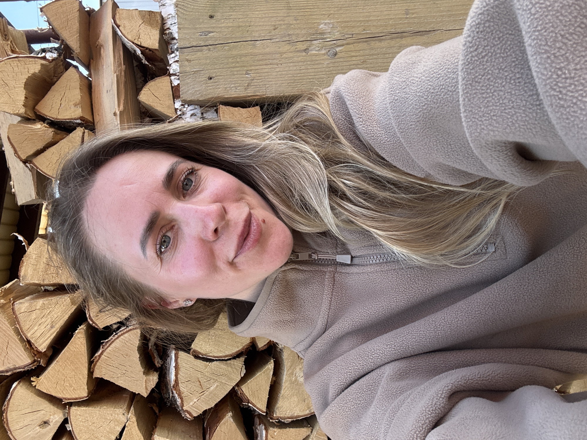 A woman is posing for a selfie outdoors next to a stack of firewood. She is wearing a light brown fleece jacket with a zipper. Her hair is blond and wavy. The firewood is arranged in a haphazard stack. Behind the firewood are some weathered wooden planks. The location is not provided in the image.
