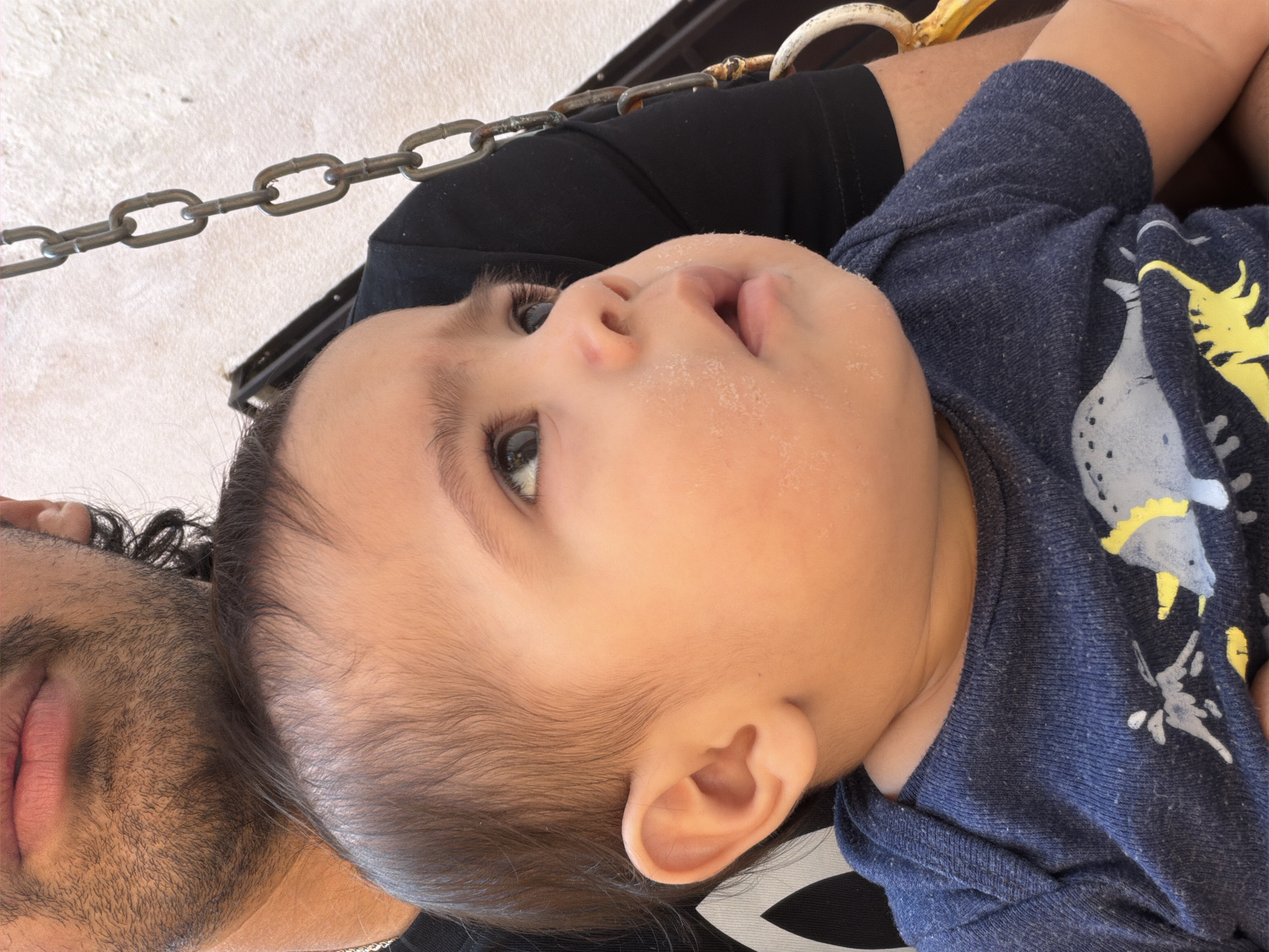 The image depicts a close-up of a baby held in someone's arms, possibly on a swing given the presence of chains. The baby is fair-skinned with dark hair and eyes, looking upwards. The baby is wearing a navy blue shirt with a yellow and white pattern. Part of the adult's face is visible, showing facial hair and lips. The adult is wearing a black t-shirt. The location, Reforma, Mexico, is not discernible from the visual information alone.