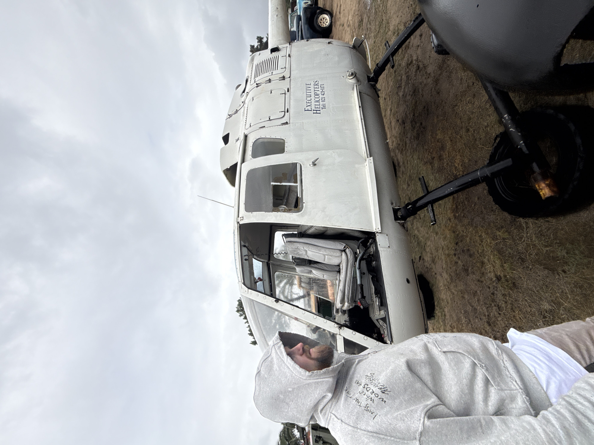 A man wearing a hooded sweatshirt is standing next to a helicopter on a grassy field. The helicopter is white with the words "EXECUTIVE HELICOPTERS" printed on the side, and the registration number "Tel: 021 421-1171". The man is looking up at the helicopter and the location is Kraaifontein, South Africa.