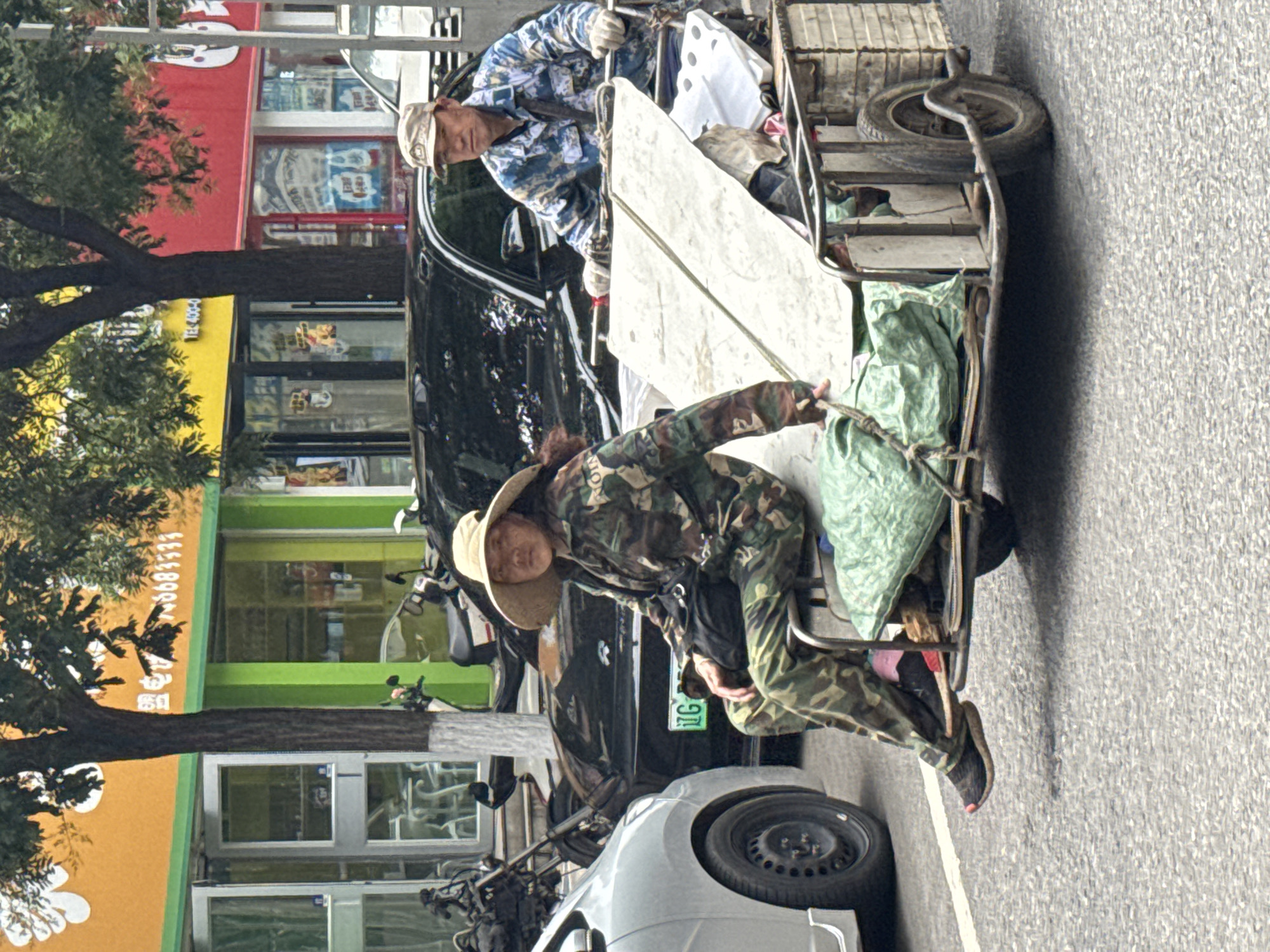 A woman wearing camouflage clothing and a wide-brimmed hat sits on a trailer being pulled by a motorcycle. She is positioned on the left side of the trailer, facing forward. To her right, a man, also in camouflage clothing and a baseball cap, operates the motorcycle. They are on a paved road in the city of Linghe, China, with shops and trees visible in the background. The trailer is loaded with various items, including green fabric bags and what appears to be sheet metal. A silver car is parked on the left side of the frame.