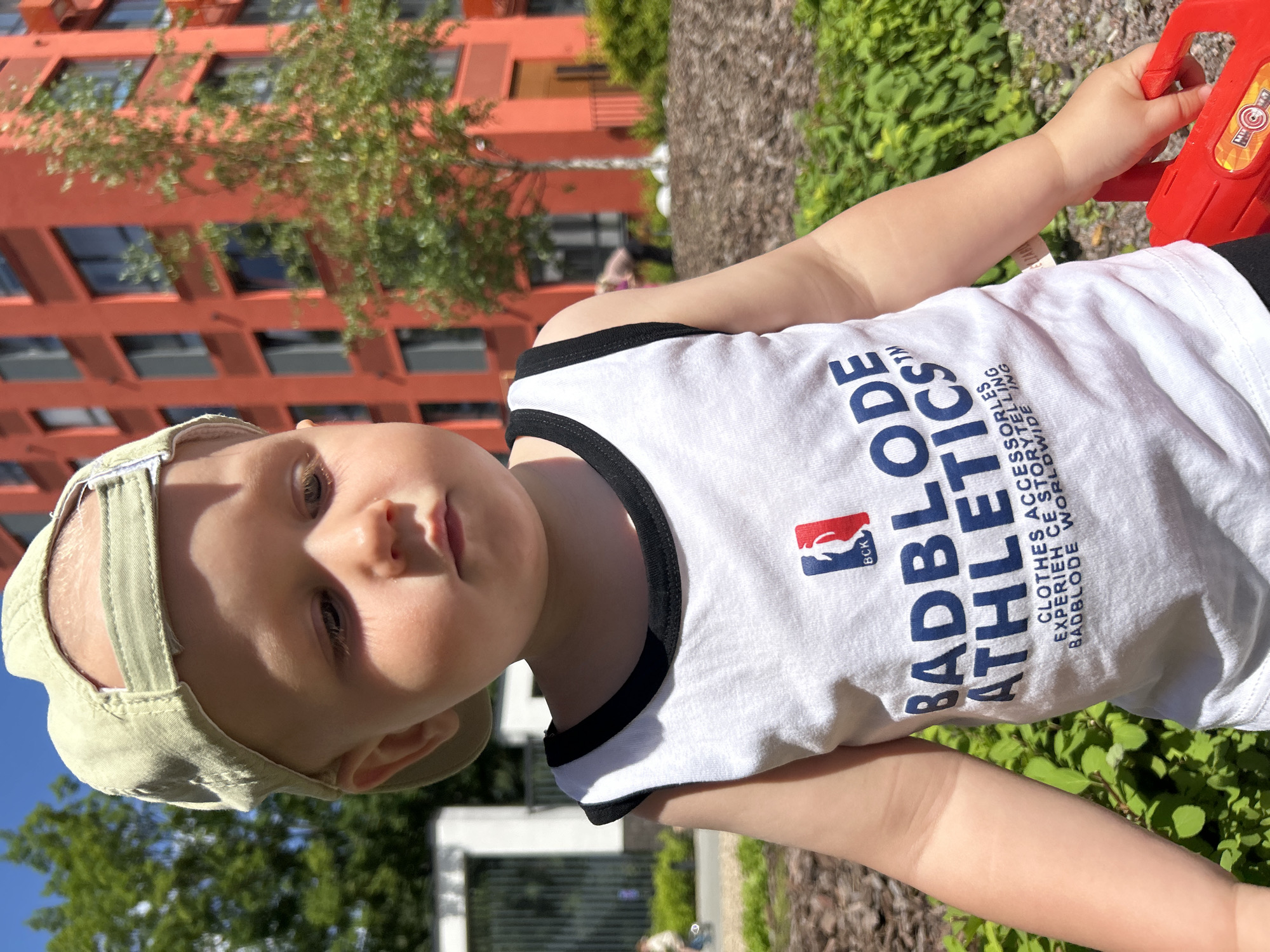 A young child, wearing a light-colored baseball cap and a white tank top with blue and black trim, is the subject of this medium shot. The child's gaze is directed downwards, to the right. The tank top features a graphic logo with the text "BADBLODE ATHLETICS" in blue. Below this, smaller text reads "CLOTHES ACCESSORIES EXPERIENCES STORYTELLING BADBLODE WORLDWIDE". The child's right hand is holding a red toy.
The background suggests an urban outdoor environment. To the left, a low, white building with dark rectangular openings is visible. Further back, a large, orange building with multiple windows stands prominently. A tree with green foliage grows in front of the orange building. The ground in the foreground is covered with mulch and green foliage. Sunlight illuminates the scene, creating shadows. The overall impression is that of a sunny day in a developed urban area, possibly a park or a courtyard within a city. There are no specific visual elements that definitively place this scene in Kaluga, Russia, beyond the user's prompt.