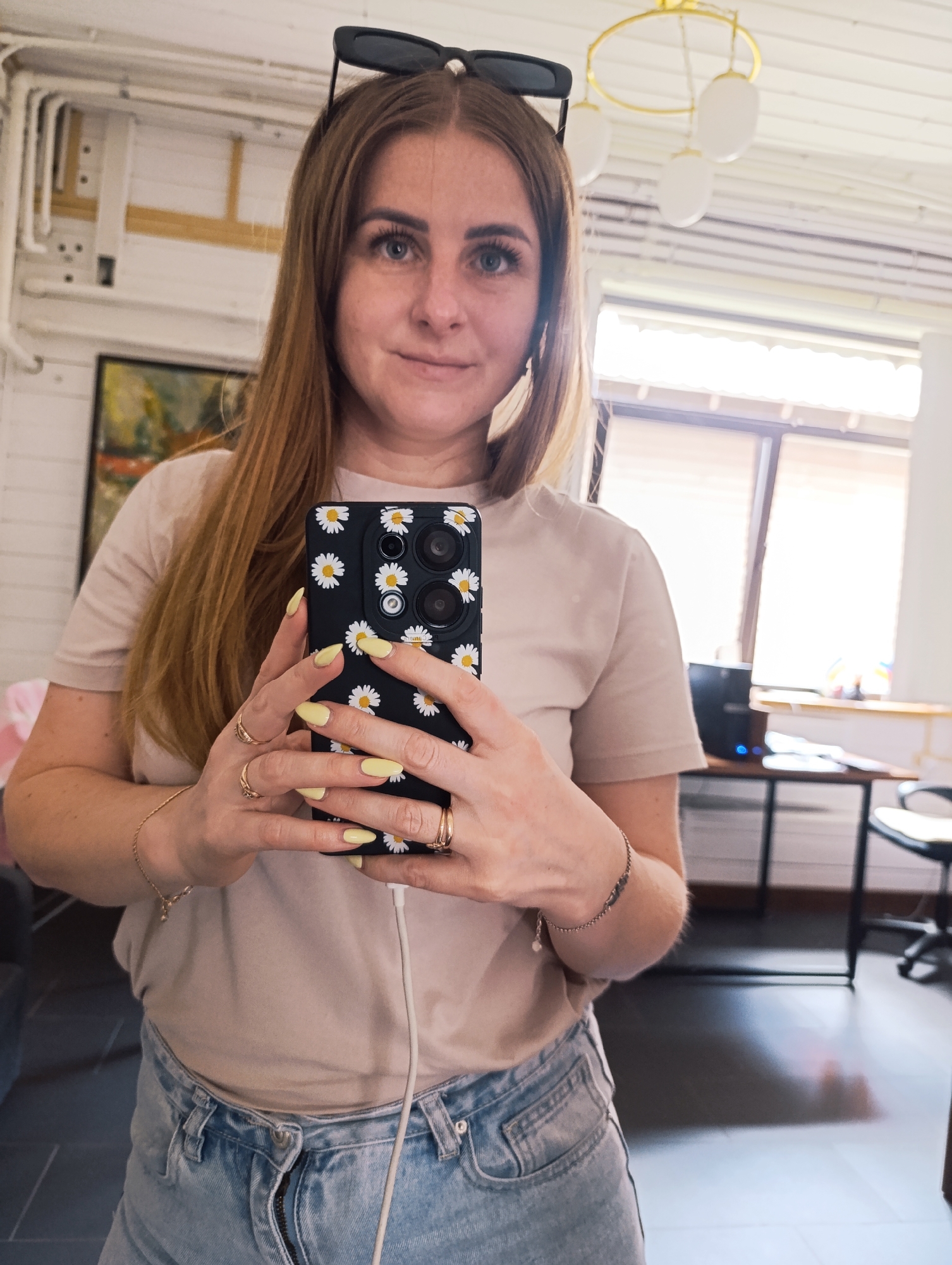 A woman is taking a selfie in an indoor setting, location unknown. She is holding a black smartphone with a daisy design on the case. She is wearing blue jeans, a beige t-shirt, and has sunglasses perched on her head. Her nails are painted a light yellow color, and she is wearing several rings. The room appears to be a home office or living space with a desk, chair, painting on the wall, and a unique ceiling light fixture. A charging cable is plugged into the phone.
