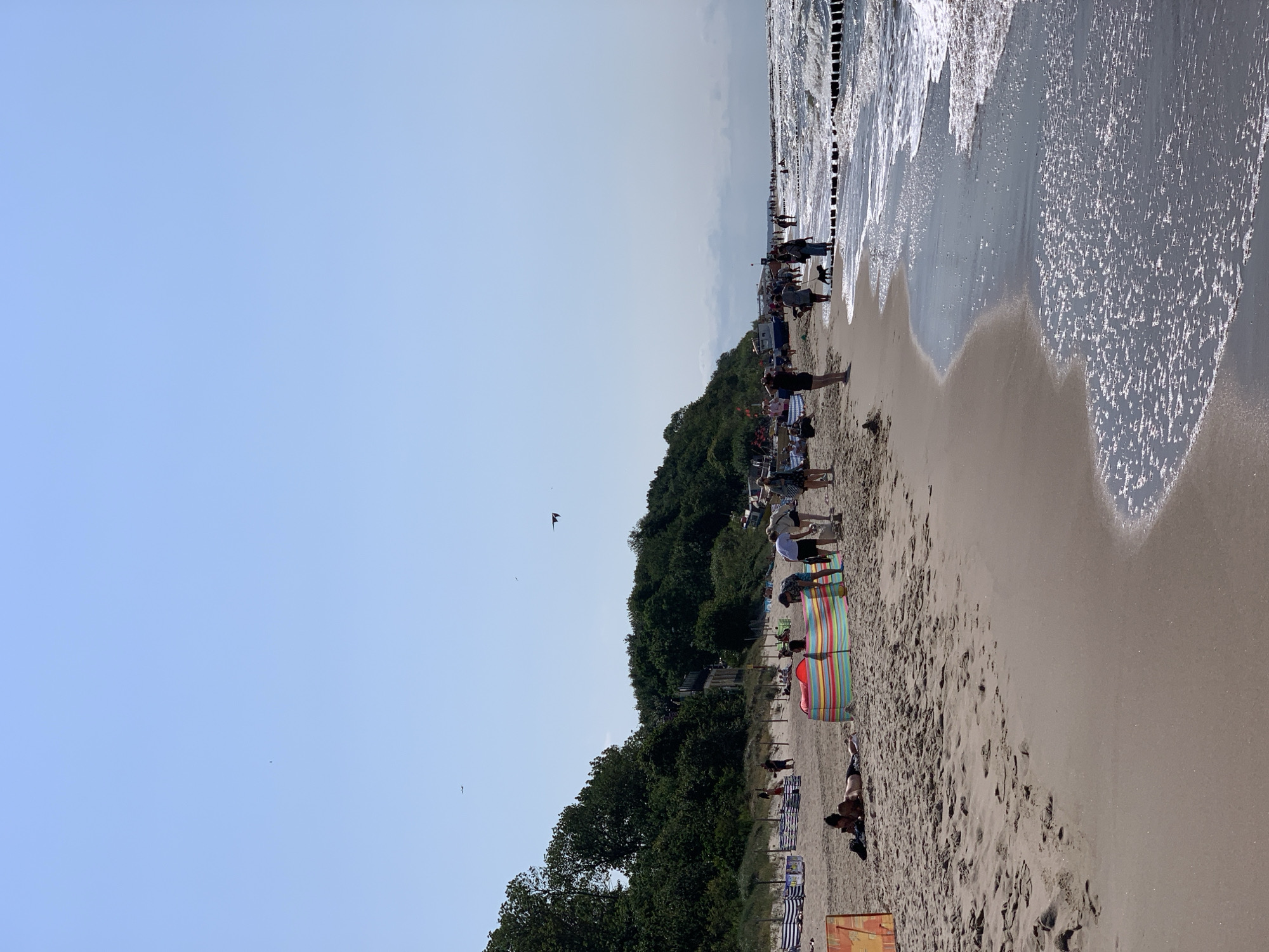 This is a photograph of a beach in Rewal, Poland, on a sunny day with a clear blue sky. The beach is populated with people enjoying the weather and the sea. In the foreground, the sand meets the water, with gentle waves washing ashore. Further up the beach, several beachgoers are visible. Some are lounging on towels, while others are standing or walking. There are beach screens set up to provide shelter from the sun. A colorful striped beach screen is particularly prominent. In the background, a line of trees and vegetation forms a natural barrier along the edge of the beach. Several small boats are visible on the sand near the tree line. A wooden breakwater extends into the sea. Two birds are flying in the sky.