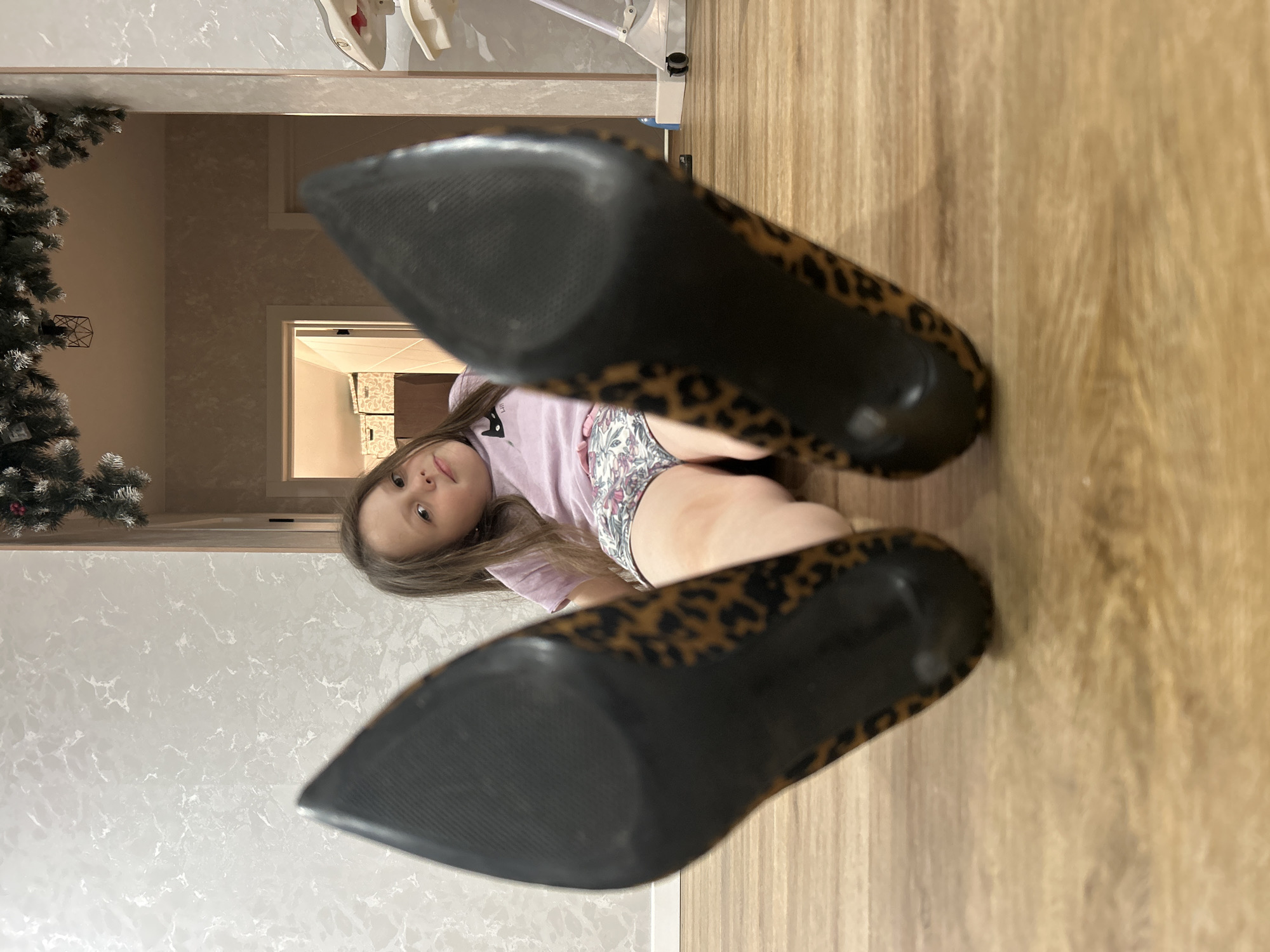 The image captures a young girl lying on a wooden floor, looking directly at the camera. She has long brown hair and is wearing a light purple t-shirt and patterned shorts. Her expression is neutral and curious. Prominently in the foreground, out of focus, are the soles of two leopard print high-heeled shoes, suggesting she might be playing with them or they are placed near her.
The setting is an indoor residential space. The floor is light-colored laminate or wood, and the wall on the left features light-toned, possibly marbled wallpaper. On the far left, a portion of an artificial Christmas tree, decorated with berries and pinecones, is visible, indicating the time of year might be winter or the holiday season. In the background, through a doorway, glimpses of another room with patterned wallpaper can be seen. Above the girl, on what would be the ceiling in a correct orientation, parts of a white household item, possibly a clothes drying rack, are visible.
The photograph is taken from a low perspective, creating an intimate view of the girl and the shoes. The lighting appears to be artificial and soft, illuminating the scene without harsh shadows. There is no visible text in the image. The overall impression is one of a domestic moment, perhaps a child playfully trying on adult shoes in a home in Chaikovsky, Russia.