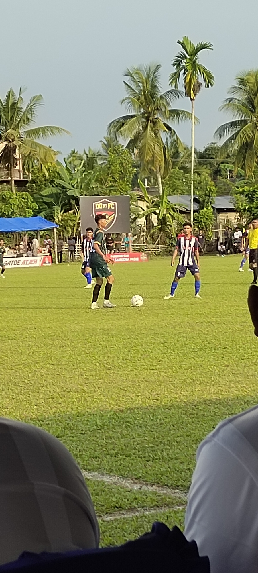 In Payateungoh, Indonesia, a football match is underway on a grassy field under a clear sky. Two male players are the central focus. One player, wearing a dark green jersey with white accents and black shorts, is positioned on the left, with his body turned slightly towards the right. He is actively engaged with a white football at his feet. The second player, in a blue and white striped jersey with blue shorts, stands on the right, facing the first player. Several other players in similar uniforms are visible in the background, along with spectators and a tent. A large banner displaying the logo "DGY FC" is prominently displayed behind the players. The surrounding environment features lush green vegetation, including palm trees, and a cluster of buildings. The foreground shows the lower bodies of spectators.