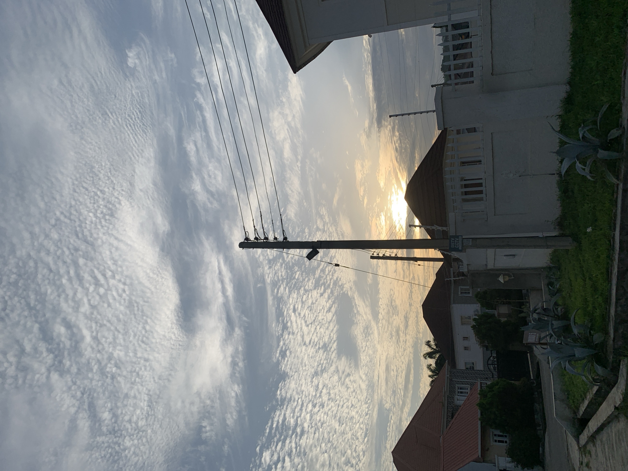 The image depicts a street scene in Abuja, Nigeria, at what appears to be dawn or dusk. The sky is filled with a pattern of cirrocumulus clouds, creating a textured, mackerel-like appearance. A low-angle view captures the sky above residential buildings. Several houses with red and brown tiled roofs are visible, along with white perimeter walls topped with fencing. Utility poles with multiple wires are prominent in the foreground and mid-ground, suggesting an active electrical grid. Vegetation, including green grass and large-leafed plants resembling agave, grows along the roadside and in front of the walls. A street sign labeled "ROAD 5" is partially visible. The overall lighting suggests a natural, ambient light source from the setting or rising sun, casting a warm glow on parts of the scene. There are no people visible in the image.
