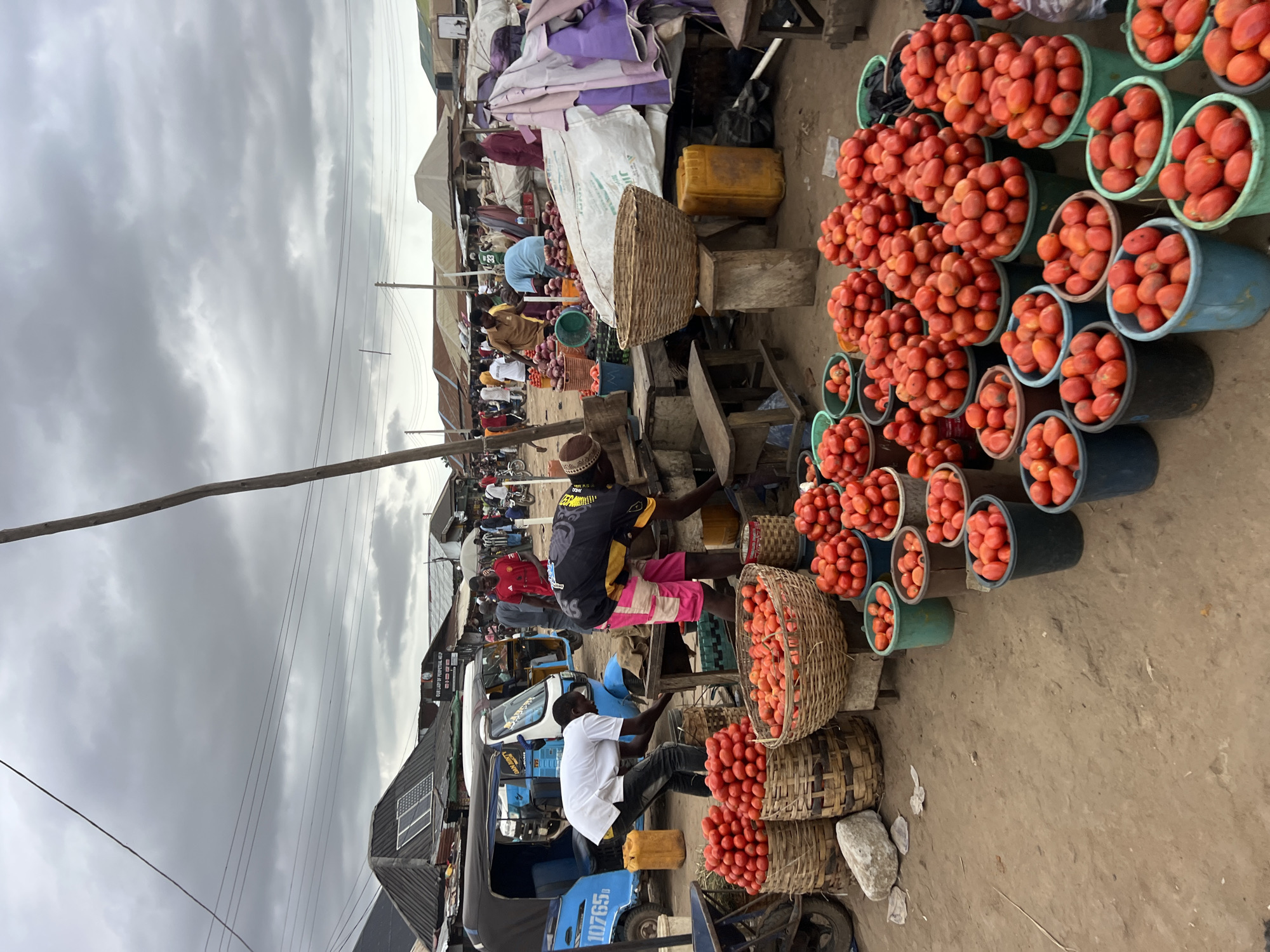 This is a photograph of an outdoor market in Oko Amakom, Nigeria, on a cloudy day. The foreground is filled with numerous buckets and baskets overflowing with ripe red tomatoes. Several people are visible in the background, engaged in activities related to the market. To the left, two men in casual attire are attending to the tomatoes. One man in a white shirt is leaning over, while another man in a black and yellow shirt with pink shorts is also working near the produce. In the middle ground, a blue and white motorized tricycle, commonly known as an "okada" or "keke napep" in Nigeria, is parked. The background reveals more market stalls, awnings, and a crowd of people. The ground is dusty earth, typical of an outdoor market. The overall scene depicts a vibrant, bustling agricultural market.
