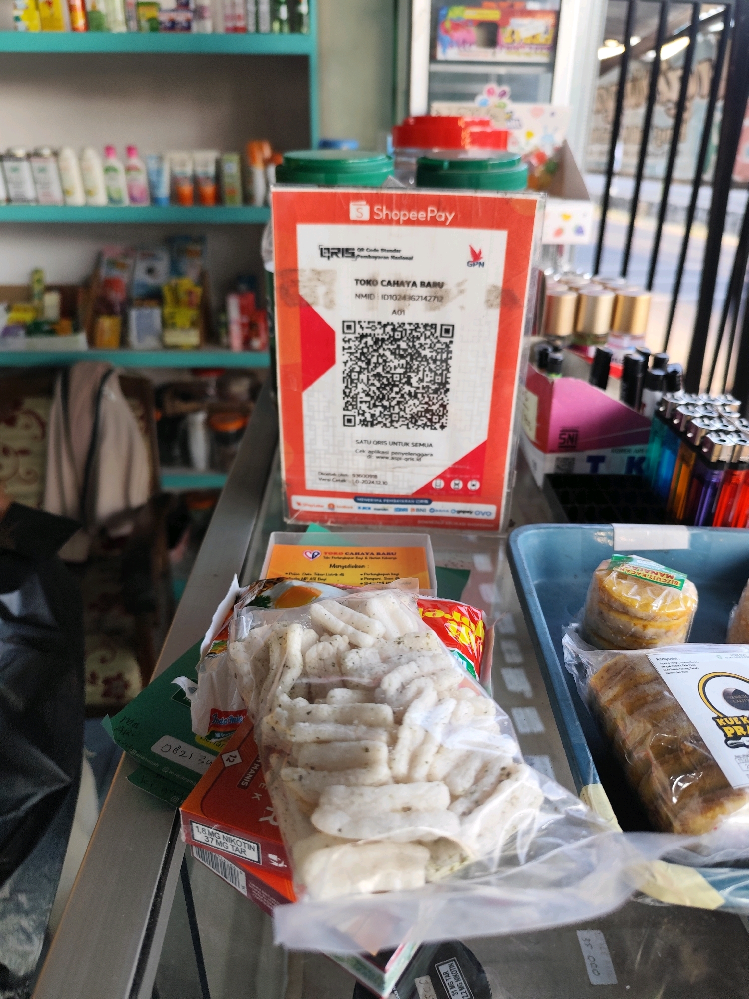 The image shows a close-up view of a counter in a small shop. On the counter, there is a ShopeePay QR code standee, a package of what appears to be snacks, and various items for sale, including lighters and some packaged goods. In the background, shelves are stocked with bottles and other products. The overall impression is of a retail environment, likely a convenience store or a small grocery shop. The items on display suggest a variety of consumer goods are available. The focus is on the QR code payment option, indicating a modern payment method being offered in this establishment.