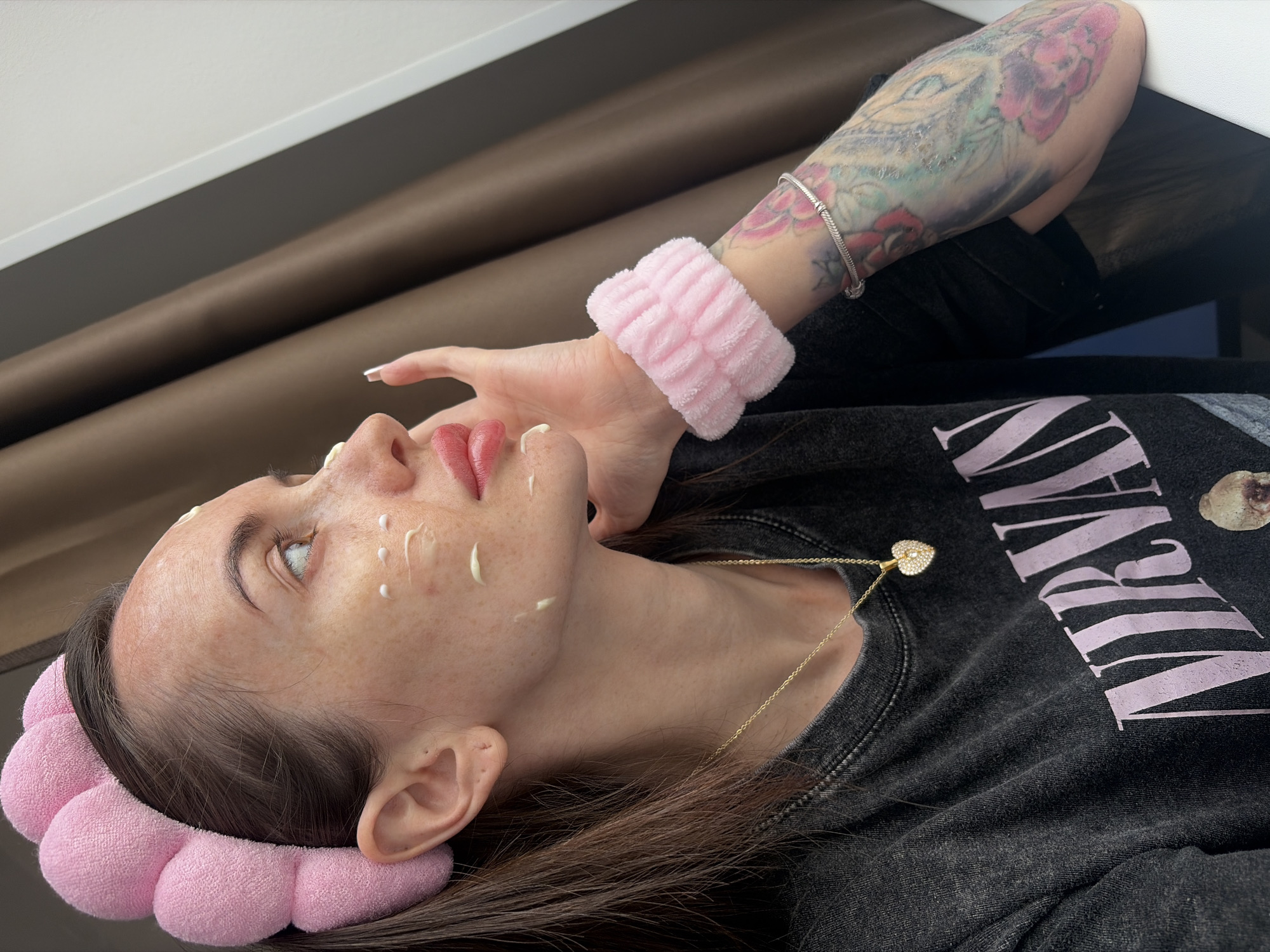 A woman is depicted lying on her back, viewed from a slightly elevated side angle. She has long, dark brown hair pulled back by a soft, pink, flower-petal-like headband. Her face, which is pale with some visible freckles, has small dots of a white or cream-colored product, likely a mask or cream, applied to her forehead, cheekbones, and beside her nose. Her eyes are open and gaze upwards, and her lips are slightly parted.
She is wearing a dark gray or black long-sleeved t-shirt with the word "NIRVANA" printed in light pink capital letters across the chest. A thin gold chain necklace with a small, heart-shaped, jeweled pendant hangs around her neck. Her left arm is visible, adorned with extensive, colorful tattoos featuring floral designs and what appears to be part of a face or eye. A fluffy, pink wristband is worn on her left wrist, and her fingernails have long, light-colored polish.
The setting appears to be indoors, with a brown fabric background, possibly curtains or furniture upholstery, and a white or light gray surface above it. The activity suggests a self-care or beauty routine, with the woman either applying or waiting for a facial product to work. Her expression appears calm and focused. The image provides no specific time of day or weather cues, but the provided context places the scene in Saint Petersburg, Russia.