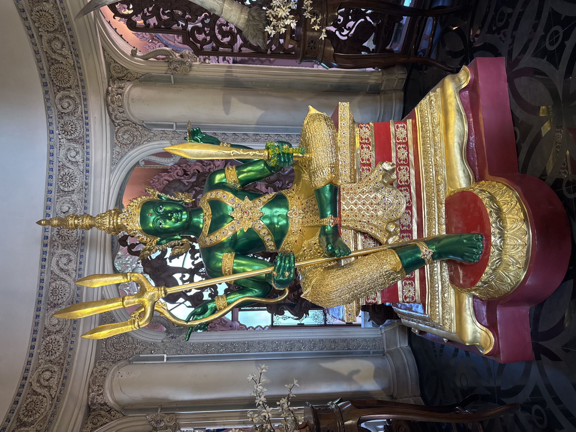 A vertically-oriented photograph features a green and gold statue seated atop a decorated pedestal. The statue, with its green skin, is adorned with gold embellishments and holds a golden trident in one hand and a golden sword in the other. It is seated on a raised, red and gold decorated throne. The statue is prominently displayed in a room with ornate white walls, with detailed carvings and floral designs. The floor appears to be tiled with dark geometric patterns. The location is the city of Samrong Nuea Subdistrict Municipality, Thailand.