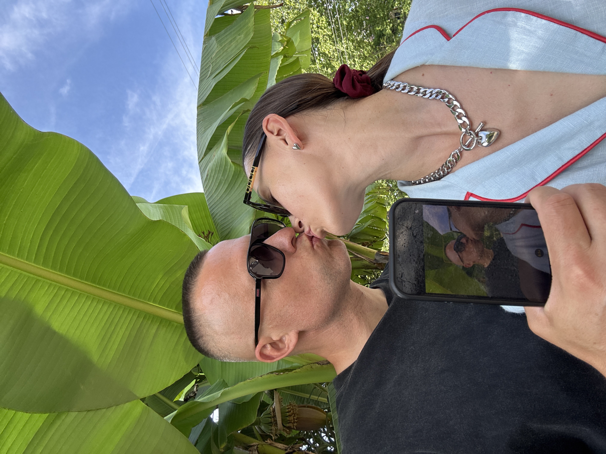 A man and a woman are kissing in front of large banana leaves and a blue sky with clouds. The man is wearing a black t-shirt and sunglasses. The woman is wearing a light blue jacket with red trim, sunglasses, and a silver chain necklace with a heart pendant. She has a burgundy scrunchie in her hair. The man is holding a smartphone, which displays a reflection of them kissing. The scene appears to be outdoors in a tropical or subtropical environment. There are no visible landmarks that specifically place the location in Sochi, Russia.