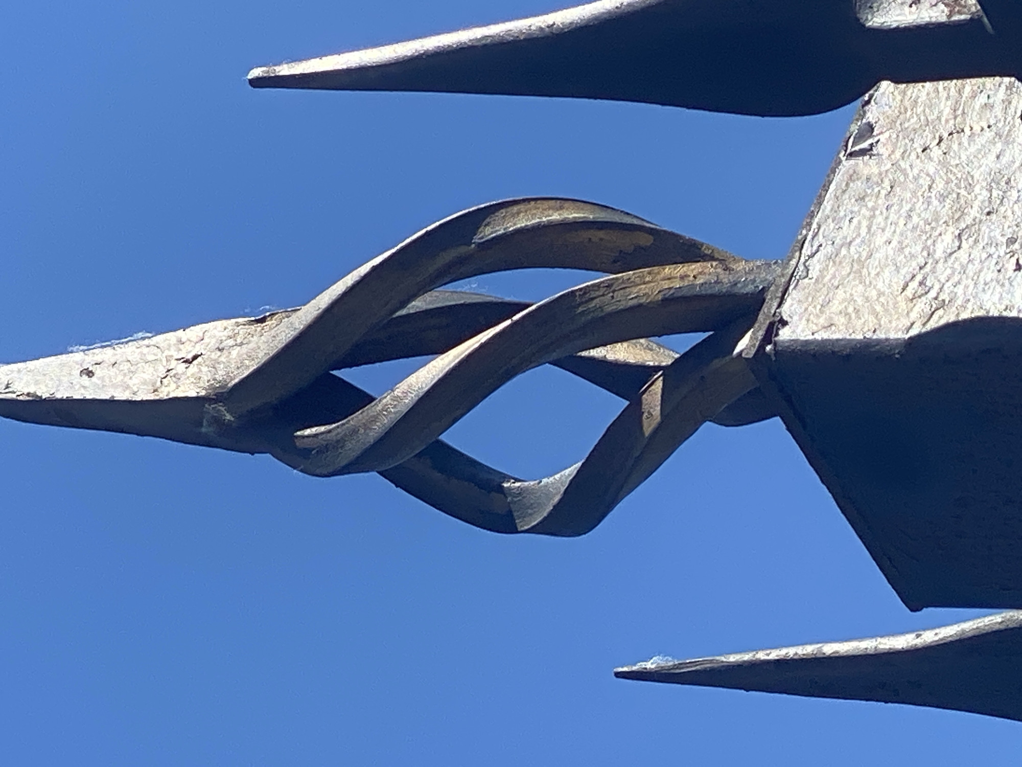This is a close-up, low-angle, outdoor photograph of a decorative metal finial against a clear blue sky. The finial is part of what appears to be a fence or gate, with pointed spikes visible on the left and right edges of the frame. The central element is an ornate, twisted metal design that resembles a stylized flame or double helix, made of dark, weathered metal. The surface of the metal shows signs of age and wear, with some chipping and a lighter, metallic sheen in places where the darker coating has worn off. The base of the finial is a textured, square stone or concrete block, also showing wear and tear. There are no people or animals visible in the image. The lighting suggests a sunny day. This image is not associated with Novokuznetsk, Russia; it depicts a decorative architectural element, likely found in various urban or historical settings.
