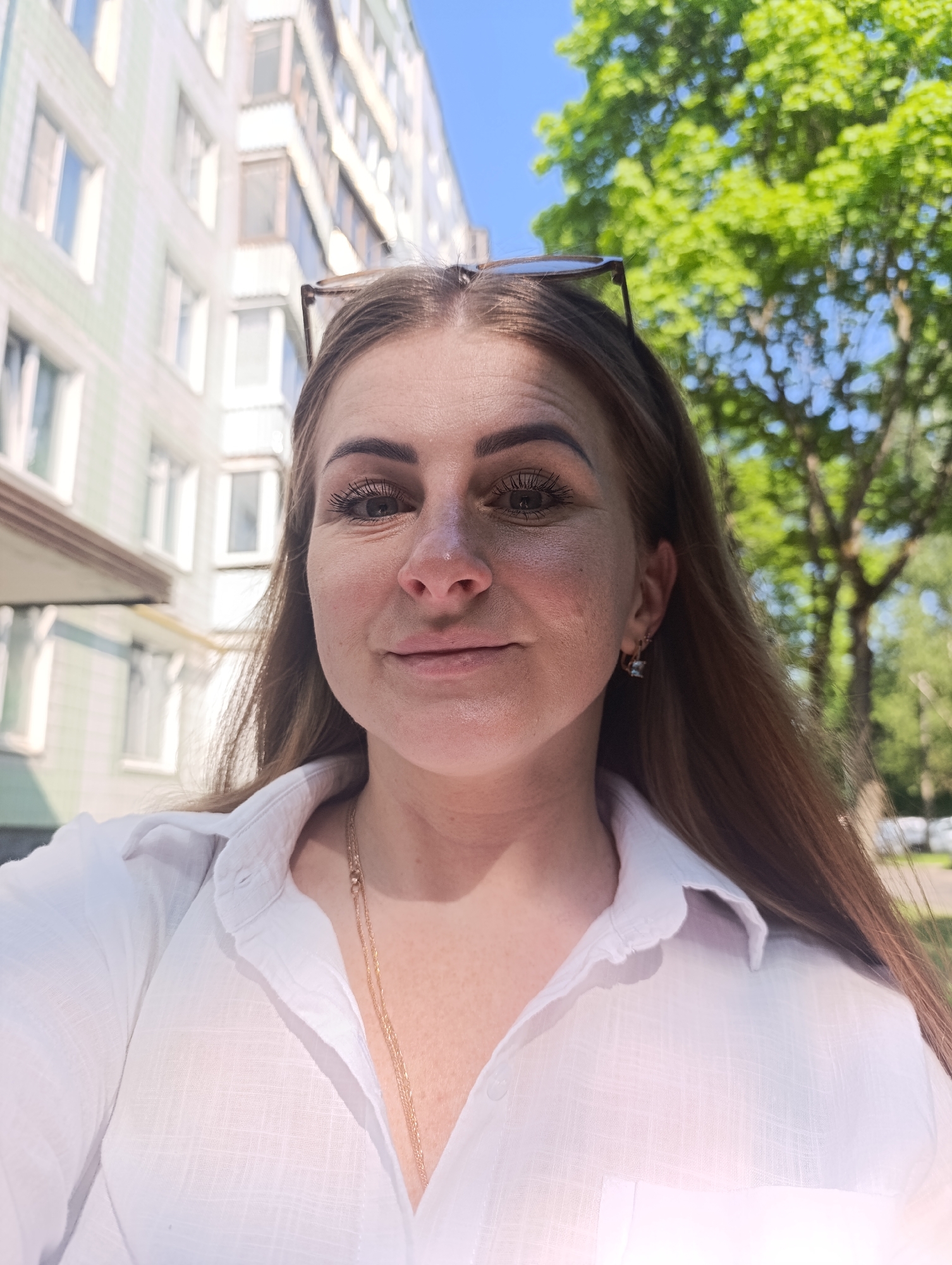 A woman is taking a selfie outdoors in the city of Unknown, Unknown. She is wearing a white button-down shirt and a gold chain necklace. Sunglasses are perched on top of her head. Behind her is a multi-story building with light-colored facades and numerous windows. To her right are green trees and vegetation. The scene appears to be daytime.
