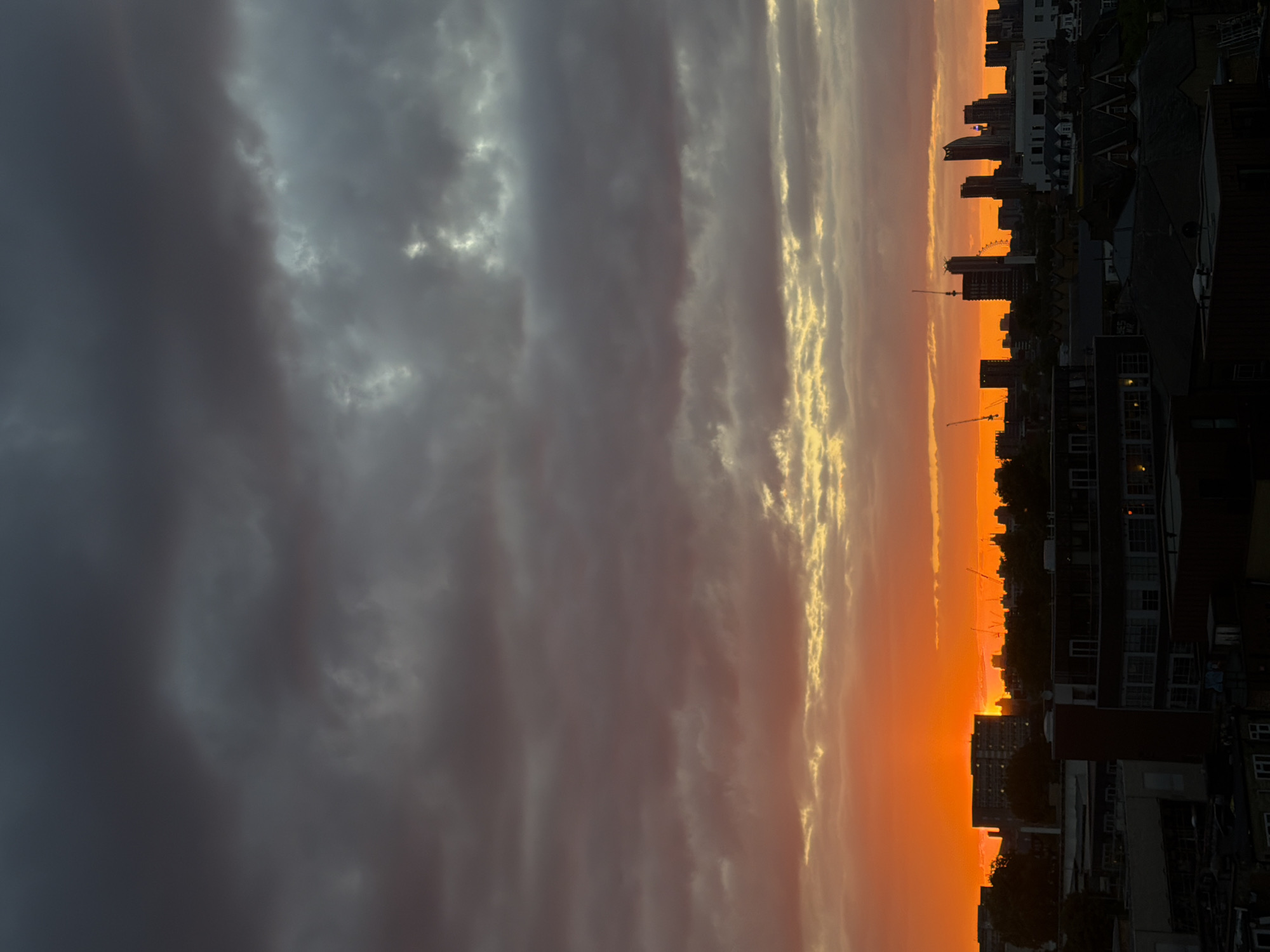 The image captures a vibrant sunset over London, United Kingdom. The sky is a dramatic blend of dark, stormy clouds on the left transitioning to fiery orange and yellow hues on the right where the sun is setting. The silhouette of the London Eye is visible among other buildings and skyscrapers in the city's skyline. Closer to the foreground, the rooftops of buildings are visible, some with visible windows and architectural details. Overall, the image presents a mix of ominous weather and the beauty of a sunset over a major urban landscape.