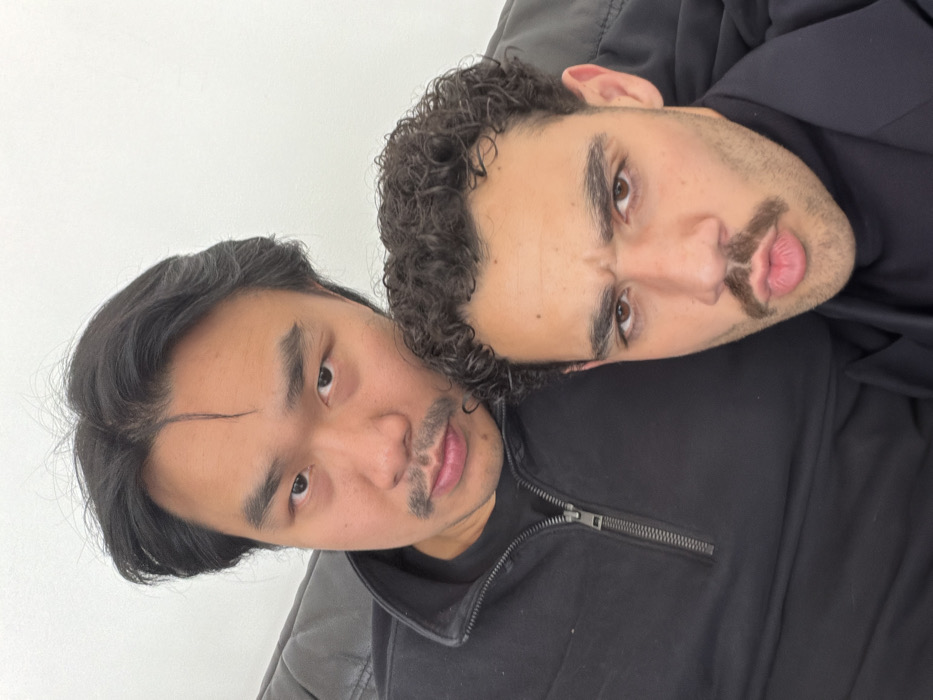The image displays two adult males positioned in a close-up, indoor selfie. The individual in the background, appearing to be of East Asian descent, has dark, medium-length hair and a thin mustache. He wears a black quarter-zip garment and maintains a neutral expression while looking directly at the camera. The individual in the foreground, appearing to be of Mediterranean or similar descent, has dark, curly hair and a prominent mustache. He wears a dark turtleneck sweater under a dark blazer, with his lips pursed and eyebrows furrowed, also looking towards the camera. The background consists of a plain white wall and a portion of a dark sofa. There are no discernible visual cues within the image to identify its specific location as Paris, France.