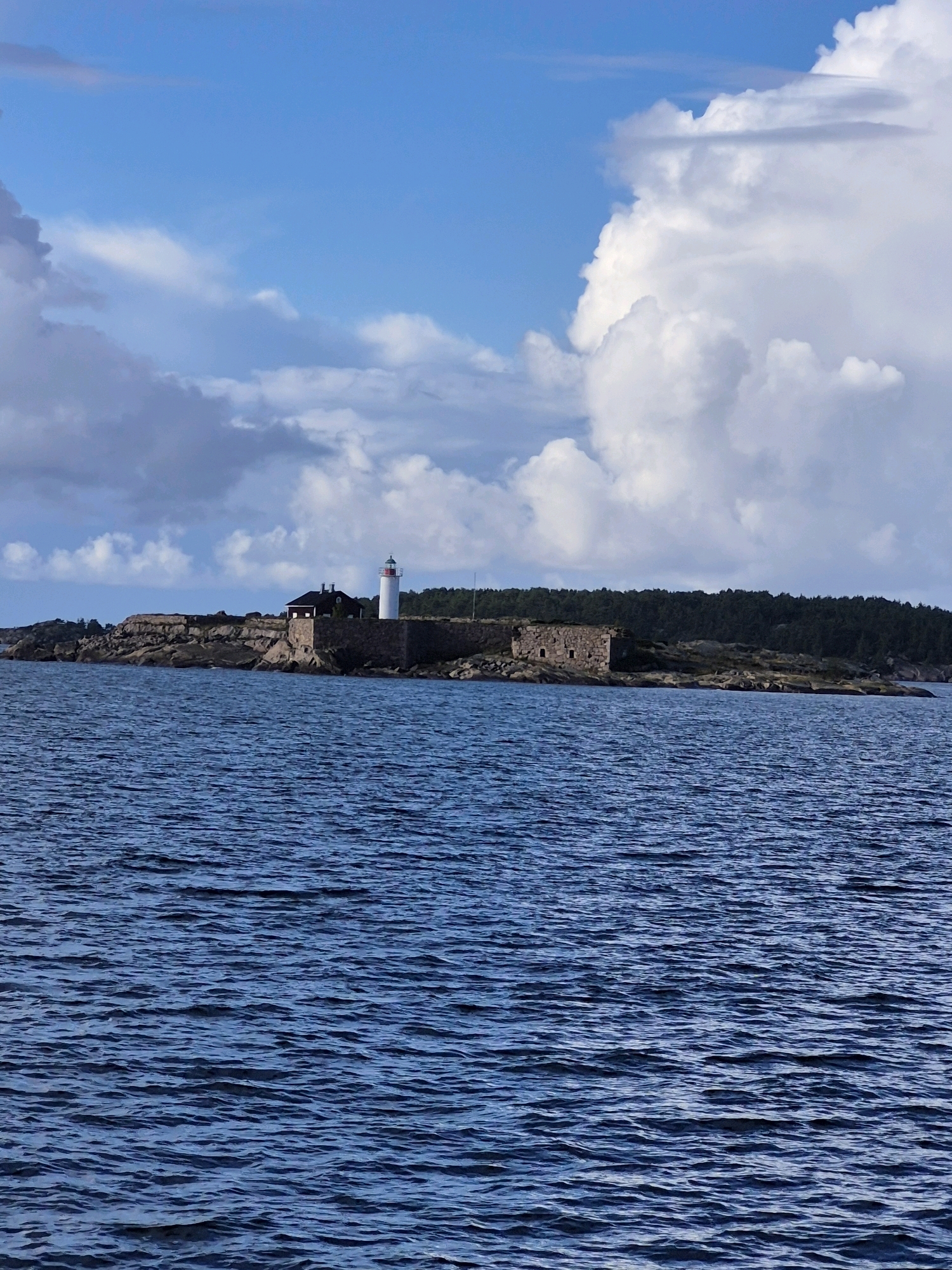 The image depicts the Hanko Lighthouse, also known as the Gustavsvärn Lighthouse, located on the island of Gustavsvärn in Hanko, Finland. The lighthouse, a white tower with a red top, stands prominently on a rocky island. Adjacent to the lighthouse is a building with a dark roof, likely part of the lighthouse keeper's quarters. The island is surrounded by stone fortifications, suggesting a historical defense structure. The scene is set against a backdrop of a deep blue, rippling sea in the foreground and a lush, dark green forest lining the horizon behind the island. The sky is a mixture of bright blue with dramatic, fluffy white clouds. There are no people visible in the image. The overall atmosphere is serene and somewhat historical, capturing a coastal landmark.