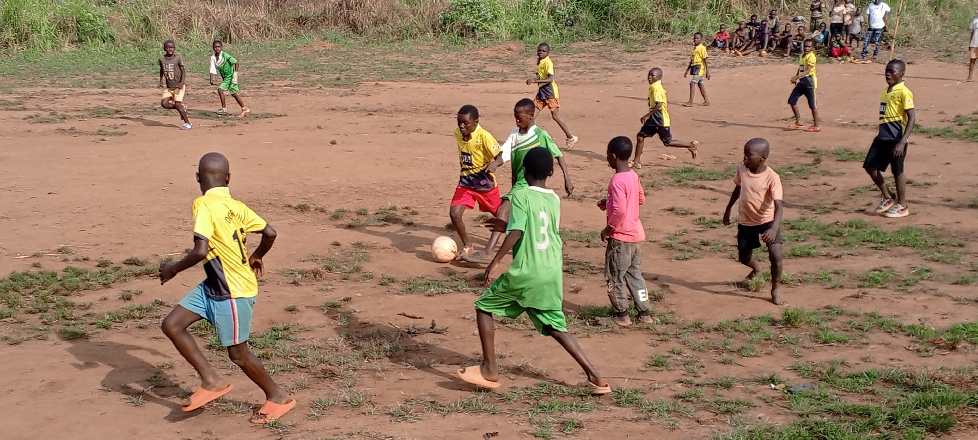 In Ngalula, Democratic Republic of the Congo, a soccer game is in progress on a dirt field. There are two teams of young boys, one in yellow and blue jerseys and the other in green and white jerseys. A soccer ball is at the center of the playing field. Other children are watching from the sidelines.