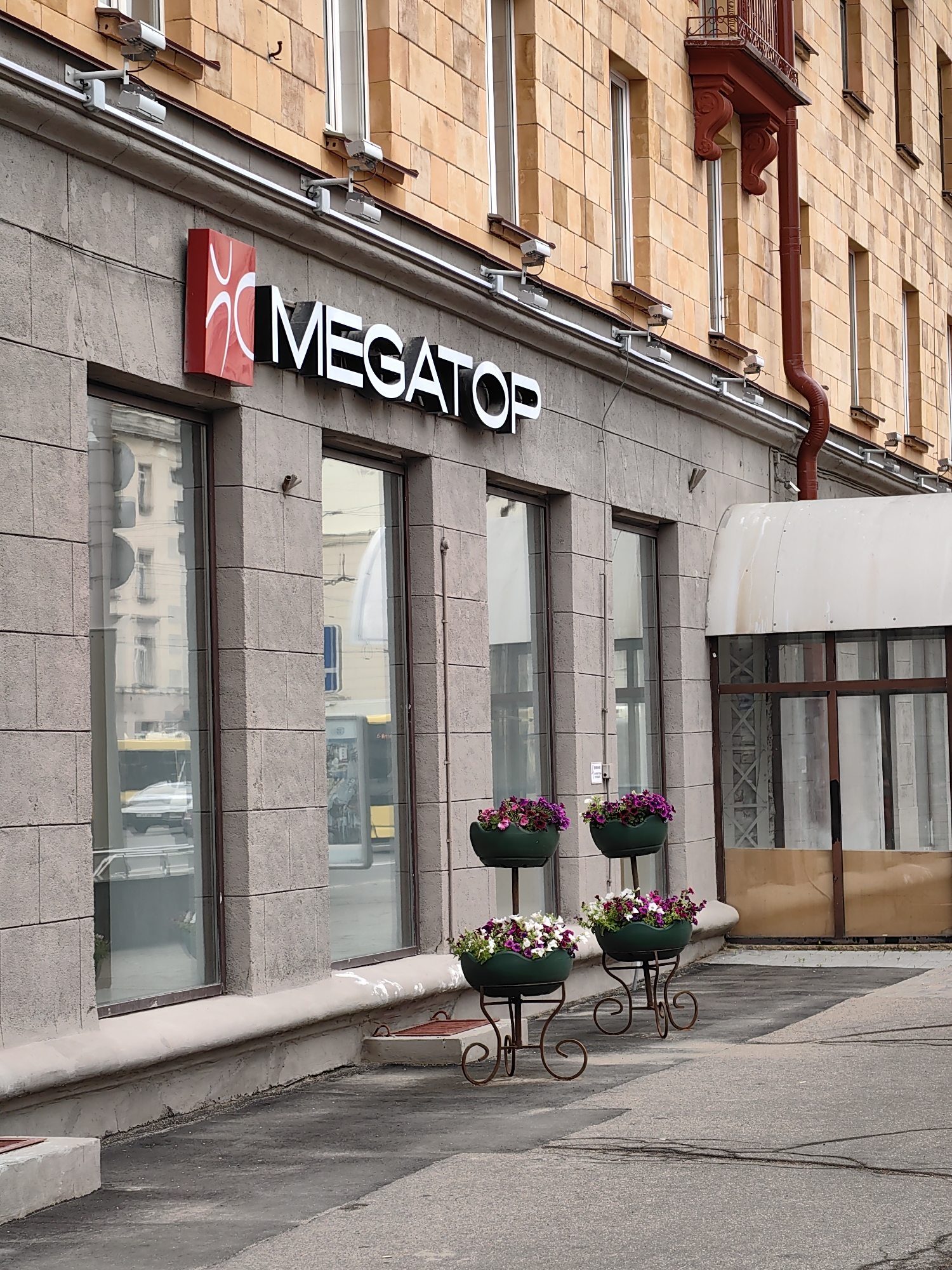 The image shows the exterior of a storefront, likely in Minsk, Belarus, based on the provided context. The building facade is constructed from light-colored stone blocks. Above the storefront windows, a sign reads "MEGATOP" in large white letters with a red rectangular logo featuring a stylized white design. Two security cameras are mounted on the facade above the sign. In front of the storefront, several decorative planters filled with purple and white flowers are placed on ornate stands. The storefront windows reflect the opposite side of the street, showing buildings and a yellow bus. To the right, there is an entrance with a white awning and a brown frame around glass doors. The ground in front of the building is paved with asphalt.
