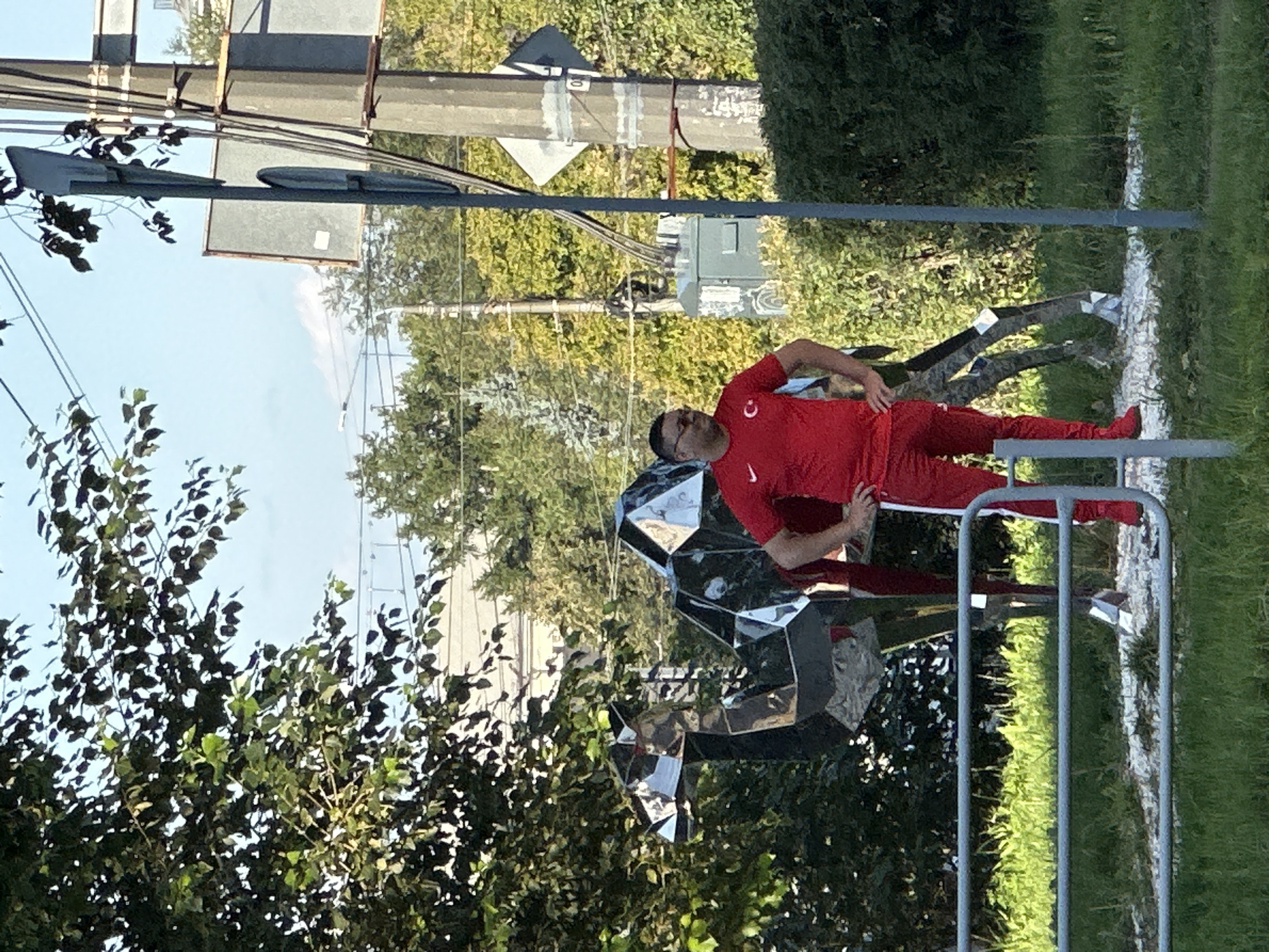 A man wearing a red t-shirt with a white logo and red pants stands with his hands on his hips next to a modern, abstract sculpture of a camel. The sculpture is made of mirrored geometric panels that reflect the surroundings. The man has dark hair and is wearing glasses. He is facing the sculpture, which is positioned outdoors on a gravelly area surrounded by grass. In the background, there are trees, a utility pole with signs, and a building. The sky is clear and blue.