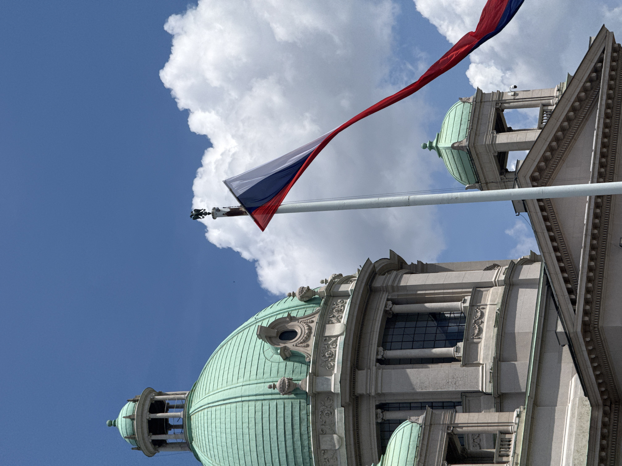 The image shows the National Assembly of Serbia building in Belgrade, Serbia, on a sunny day with a blue sky and white clouds. A large flag, appearing to be the Serbian flag with its red, blue, and white stripes, is prominently displayed on a flagpole in the foreground, fluttering in the wind. The building itself is characterized by its grand architecture, featuring domes, columns, and ornate detailing. The architectural style is neoclassical. The flag's colors are red, blue, and white. The flag is angled diagonally across the frame, with the red stripe appearing at the bottom and the blue and white stripes above it. The flag is partially obscured by the flagpole, which is white and tall. In the background, a large green dome is visible on the left side of the frame, part of the building. Another smaller green dome is visible on the right side of the frame, also part of the building. The building's facade is light-colored stone, with dark windows. The sky is a vibrant blue with fluffy white clouds scattered across it.