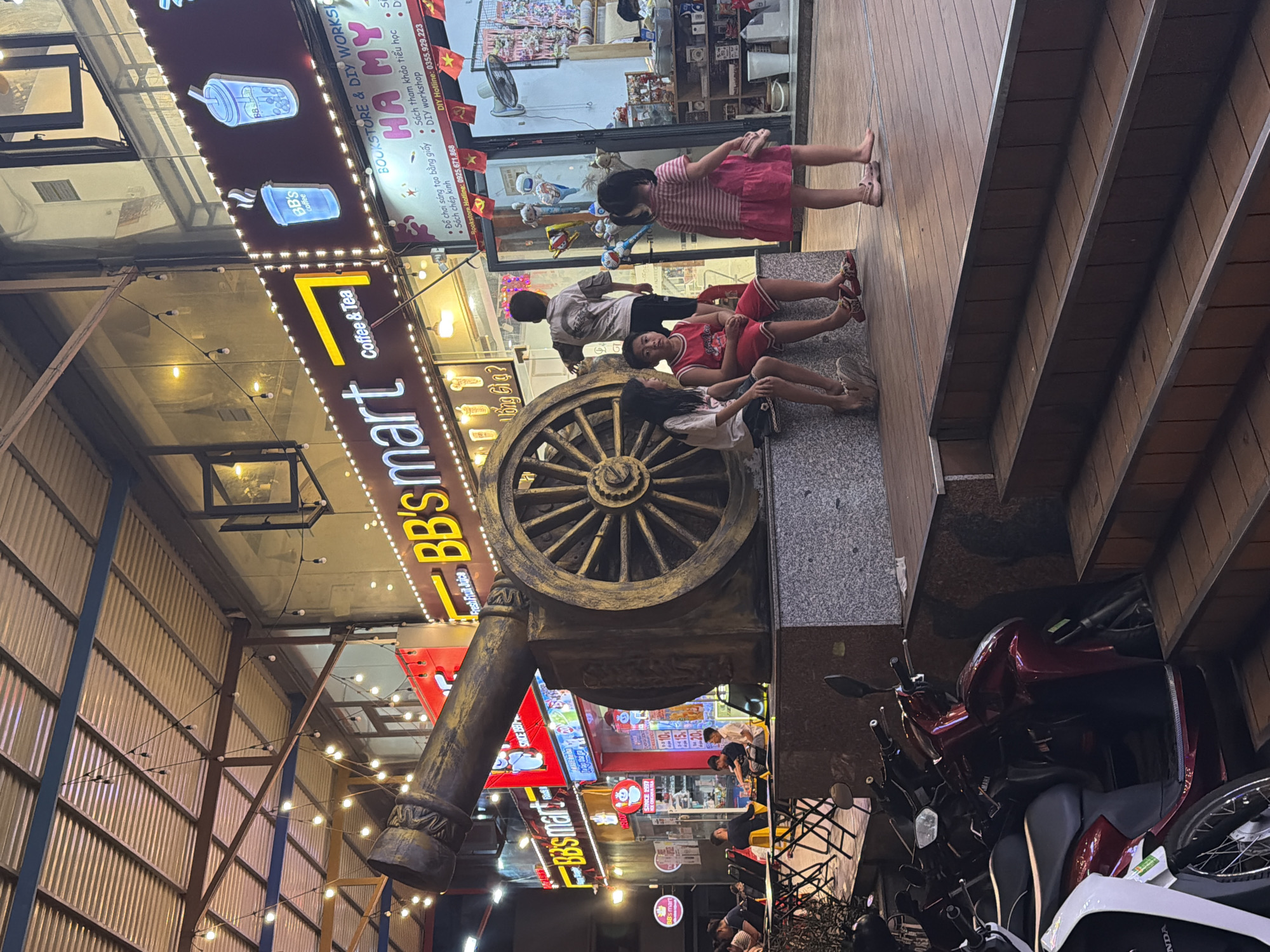 The image captures a street scene in Vietnam, likely in the Bac Nha Trang Ward, at what appears to be dusk or early evening due to the artificial lighting. The foreground is dominated by a large, ornate decorative structure resembling a cannon, with a prominent wagon wheel. To the left of this structure, a sign for "BB's mart Coffee & Tea" is brightly lit, featuring stylized images of drinks. Further left, another sign displays "BB's mart" with glowing beverages.
In the midground, three young people are casually sitting or lounging near the decorative cannon. A girl in a pink dress is standing, looking towards the left, while two others are seated. The setting seems to be a lively marketplace or shopping area, indicated by the presence of multiple illuminated signs for various businesses, including "BOOKSTORE & DIY WORKS HA MY," which lists services like "Creative Paper Crafts," "Elementary Reference Books," and "DIY Workshop," along with contact information.
In the background, more signs and shops are visible, suggesting a bustling commercial environment. The scene also includes several motorcycles parked on the right side of the image, a common sight in Vietnamese streets, and strings of decorative lights overhead. The overall atmosphere appears relaxed and social, with people enjoying a casual evening. The visible text includes business names, service offerings, and contact numbers, predominantly in Vietnamese with some English.