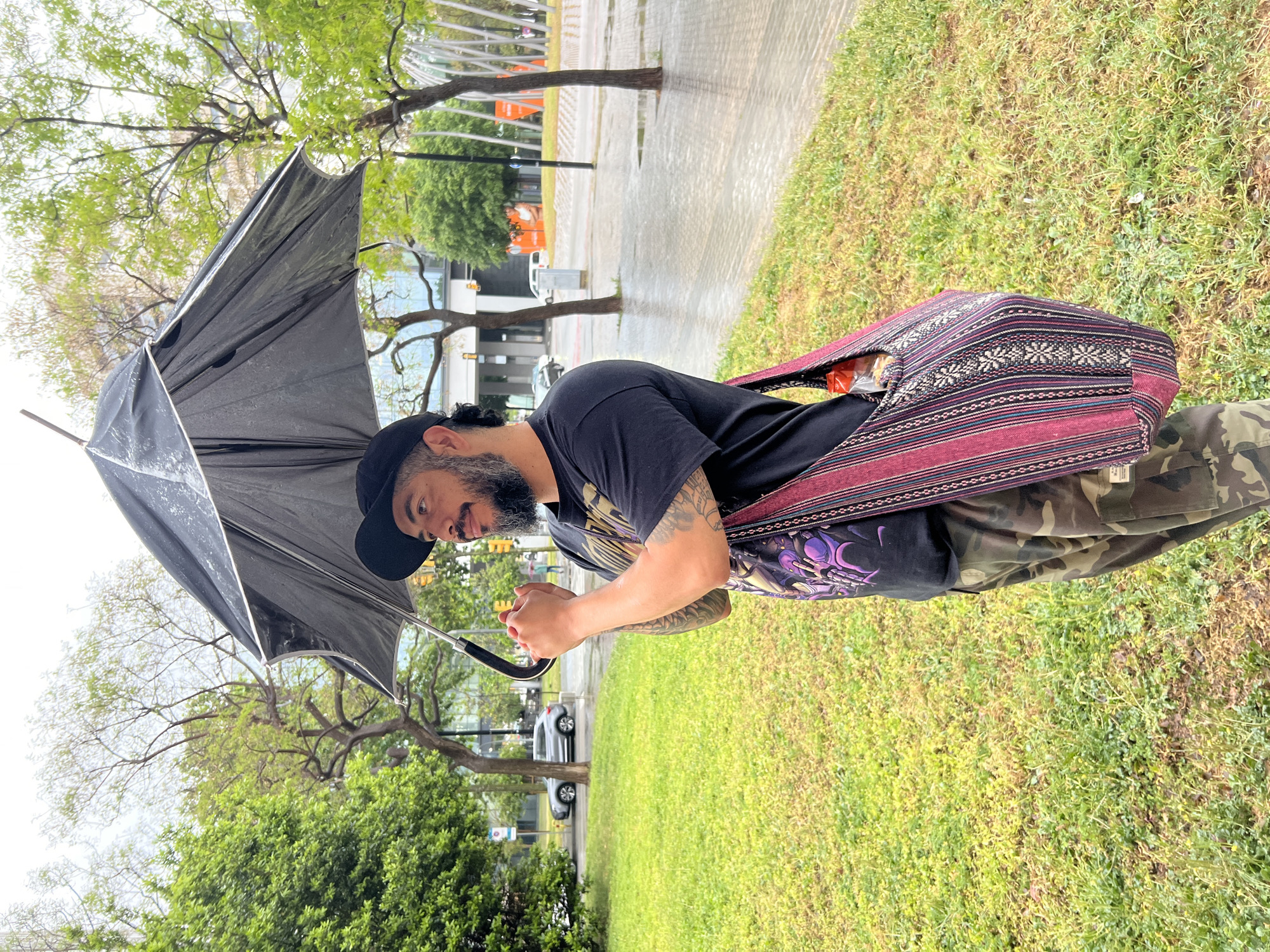 A bearded man wearing a black t-shirt, a black baseball cap, and camouflage pants is holding a black umbrella in a grassy area. He has a patterned bag slung over his shoulder. The background shows trees, a wet paved area, and buildings, indicative of an urban setting. The image was taken during a rainy day. There are no identifying landmarks in the image to confirm that the location is the city of l'Hospitalet de Llobregat, Spain.
