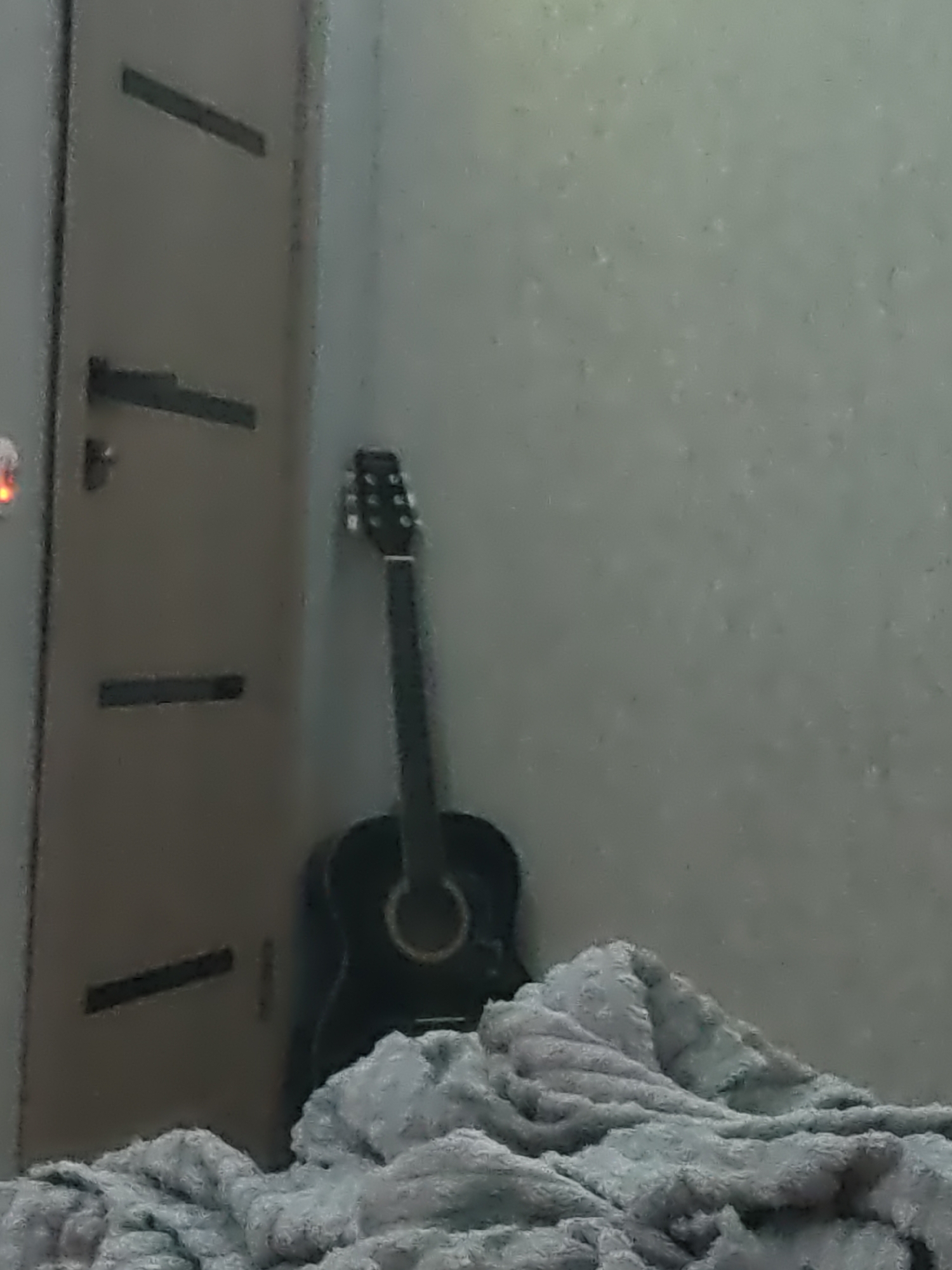 The image depicts an interior scene with a dark-colored acoustic guitar leaning against a textured, light-colored wall. To the left, a portion of a brown door or cabinet is visible, featuring horizontal dark accents. In the foreground, a plush, light-colored blanket is draped, partially obscuring the lower part of the guitar and the floor. The lighting suggests an indoor, possibly dimly lit environment. There is no indication of the specific location within the city of Kurganinsk, Russia. The scene is static and shows no human activity or interaction.