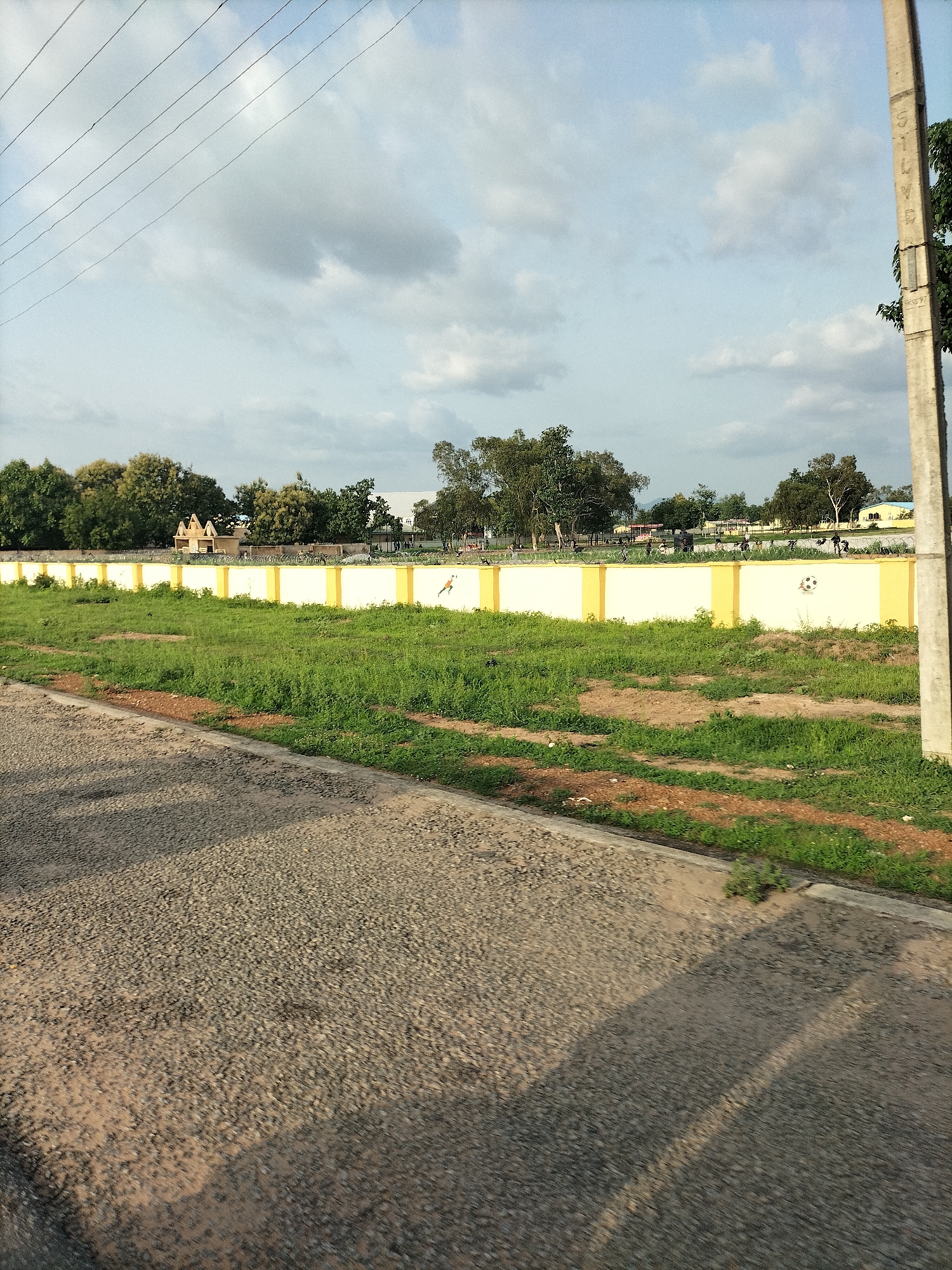 The image depicts a street-level view of a wall with yellow and white sections, bordering an area with green grass and some bare earth patches. Behind the wall, several trees and buildings are visible, suggesting an urban or semi-urban environment. A concrete utility pole stands to the right of the frame, with faint markings on it. Power lines are strung across the sky. The foreground shows a textured asphalt surface, possibly a road or pathway, with shadows cast upon it. In the background, faint figures and some bicycles can be seen along the wall. One section of the wall features a drawing of a soccer ball, and another shows a bird in flight. The overall scene appears to be outdoors in daylight, under a partly cloudy sky. This location is likely in Jalingo, Nigeria, given the architectural style and vegetation common to the region.