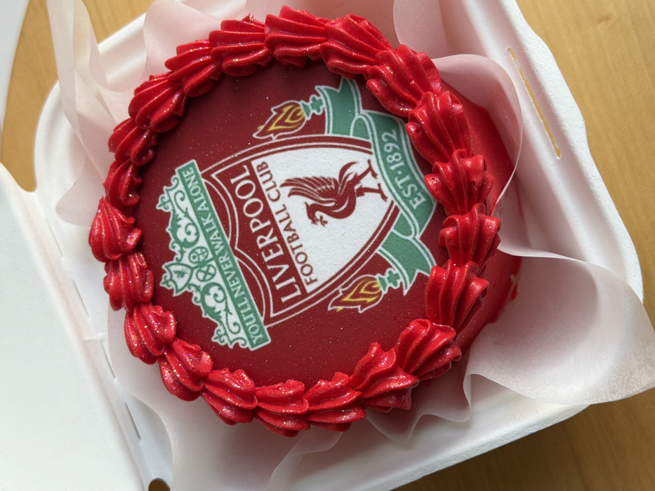 The scene is located in Tel-Aviv, Israel. A round cake, approximately 6-8 inches in diameter, is positioned within an open, white, rectangular cardboard or styrofoam takeout box. The cake's top surface is predominantly red and features a prominent edible image of the Liverpool Football Club crest. The crest is a white shield with green accents, displaying the text "LIVERPOOL FOOTBALL CLUB" and "EST·1892", centered by a red Liver bird. Above the shield, the motto "YOU'LL NEVER WALK ALONE" is written in green. Two yellow and green flames flank the lower sides of the shield. The red surface of the cake has a subtle, glittery appearance. The perimeter of the cake is decorated with a border of bright red, ruffled-patterned frosting. The cake is situated on a translucent, slightly crumpled, light pink or white paper liner inside the box. The container rests on a smooth, light brown wooden surface.