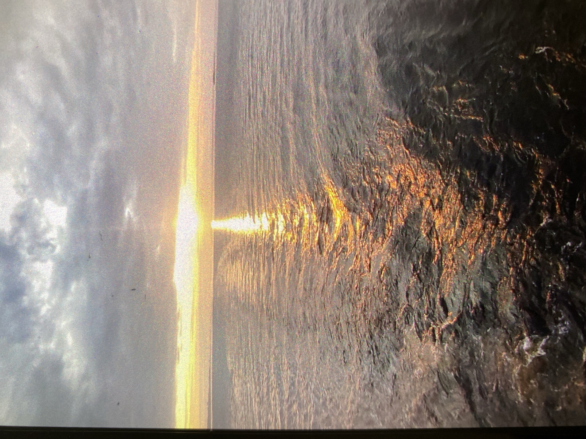 The image depicts a body of water, likely the sea, with a dramatic sunset. The sun's reflection creates a bright, vertical path of light on the water's surface. To the left, the sky is cloudy. To the right, the water appears rough and dark. Given the location of Saint Petersburg, Russia, near the Baltic Sea and the Gulf of Finland, the body of water is most likely the Gulf of Finland.