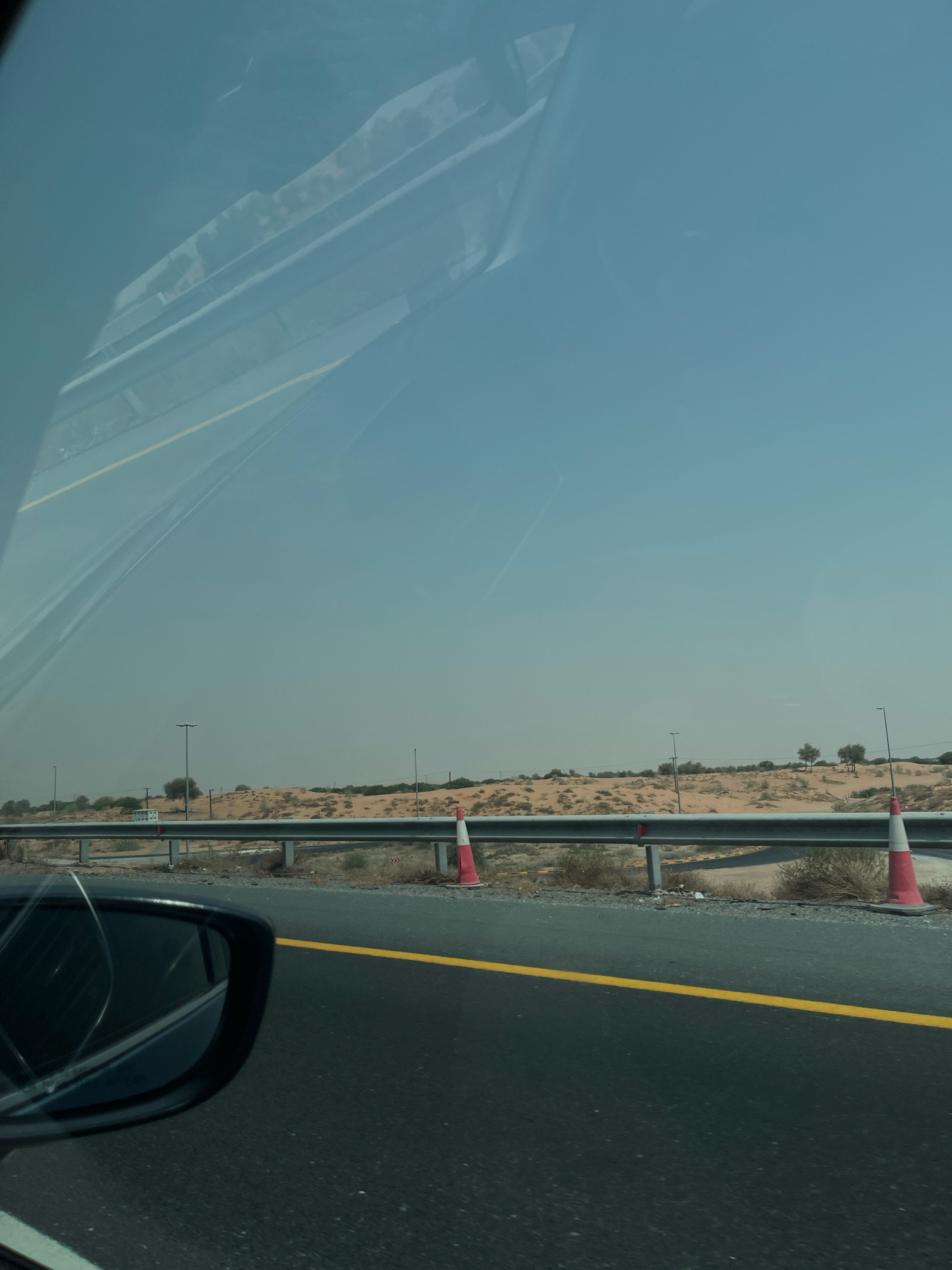 The image captures a view from a vehicle traveling on an asphalt road with a yellow line marking. In the foreground, a black side-view mirror is visible on the left. The road is bordered by a metal guardrail, beyond which lies a desert landscape characterized by sand dunes and sparse vegetation. Several traffic cones, orange and white, are positioned along the guardrail, indicating ongoing roadwork or a safety measure. Streetlights are sparsely distributed in the background. The sky is clear and blue, suggesting a daytime scene in a dry, arid environment, consistent with a location in the United Arab Emirates like Mohadhub.