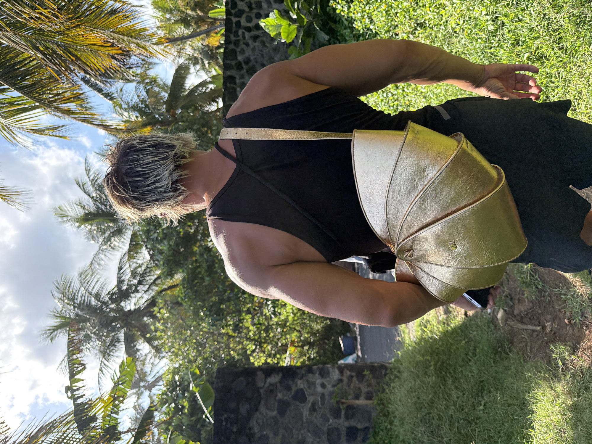 A person with fair skin, a dark tank top, and a gold-colored bag is standing in Kerambitan, Indonesia. They have blonde hair, and are holding a smartphone. The background consists of grass, trees, and a stone wall. The sky is visible with scattered clouds.