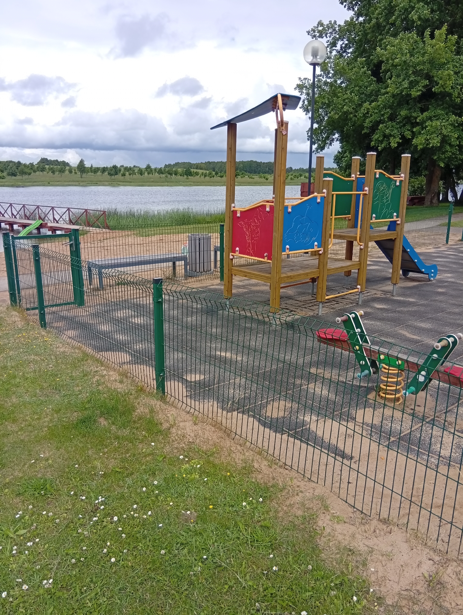 The image shows a children's playground located in Šaukėnai, Lithuania. The playground features a wooden play structure with colorful panels, a blue slide, and a seesaw with a spring mechanism. A green wire fence encloses the playground, separating it from a grassy area with small white flowers and a sandy path. In the background, a calm body of water, likely a lake, is visible, bordered by trees and vegetation. The sky is overcast with clouds. A lamppost stands near the play structure, and a trash can is situated on the playground's rubberized surface. A wooden bench is also present near the fence.
