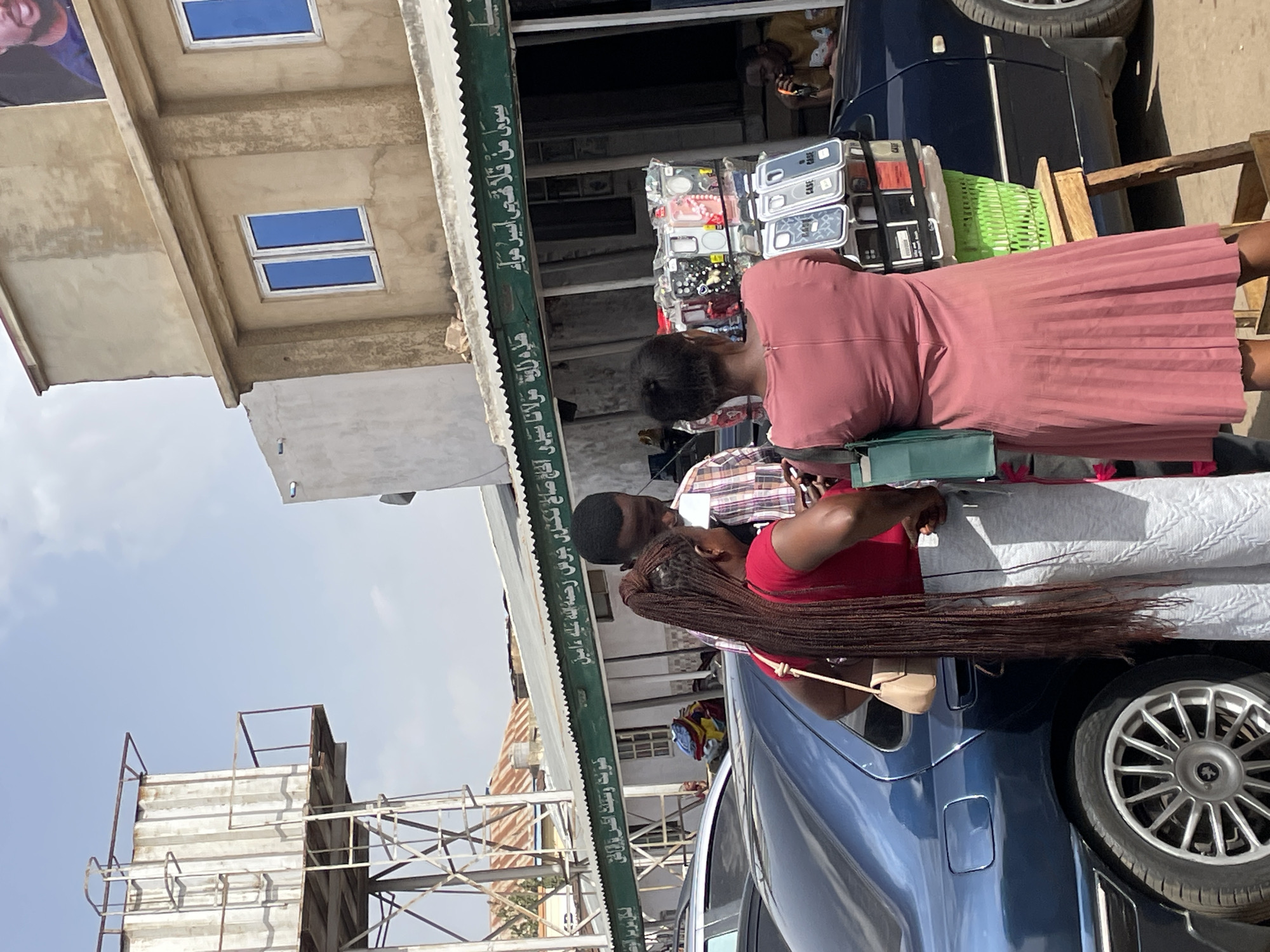 The image depicts a vibrant street scene, likely a market area, in Jos, Nigeria, under bright daytime conditions. The photograph is oriented vertically, showing the scene as if viewed from above, looking down a street or alley.
In the foreground, several individuals are present, suggesting a shopping or social interaction. A woman in a long, pink pleated dress is seen from behind, facing what appears to be a street stall. Behind her, a man in a plaid shirt is partially visible. Further to the right, another woman with long, dark braided hair, wearing a red top, is situated near a blue car, holding a light-colored purse.
The street stall features an array of mobile phone cases, neatly displayed in transparent packaging, with some visibly labeled "CASE." A bright green plastic basket rests on the stall. A dark blue car, possibly a sedan, with a silver multi-spoke wheel, is parked prominently.
The architectural background includes a multi-story concrete building with distinctive blue-framed rectangular windows on an upper floor. A large, partially visible banner featuring a man's face hangs on an upper level of the building. A green awning stretches over the stall, adorned with Arabic script. The script, identified through OCR, includes phrases such as "مولانا سيدي الحاج صالح" (Mawlana Sidi Al-Haj Saleh) and a reference to "جوس" (Jos), confirming the provided location context and suggesting a commemorative or religious inscription. The sky above is clear with scattered clouds, indicating sunny weather. Metal scaffolding or structural elements are also visible against the sky in the background, adding to the urban landscape.