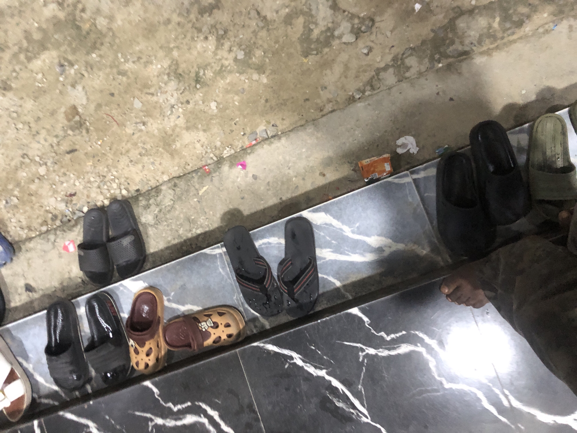 A pair of black flip-flops are resting on a tiled ledge, with their red and black straps visible. To the left of the flip-flops, a pair of tan clogs with a leopard-like pattern are positioned. Further to the left, a pair of black slip-on shoes are visible. Beyond these, a pair of brown and white sandals are partially visible at the top of the frame. A person's right foot, clad in dark pants, is visible in the lower left corner of the image, stepping onto the tiled surface. The ground next to the tiled ledge is composed of dirt and small stones, with scattered debris. The scene appears to be outdoors, possibly in an alleyway or a communal area, given the presence of various footwear and the unpaved ground.