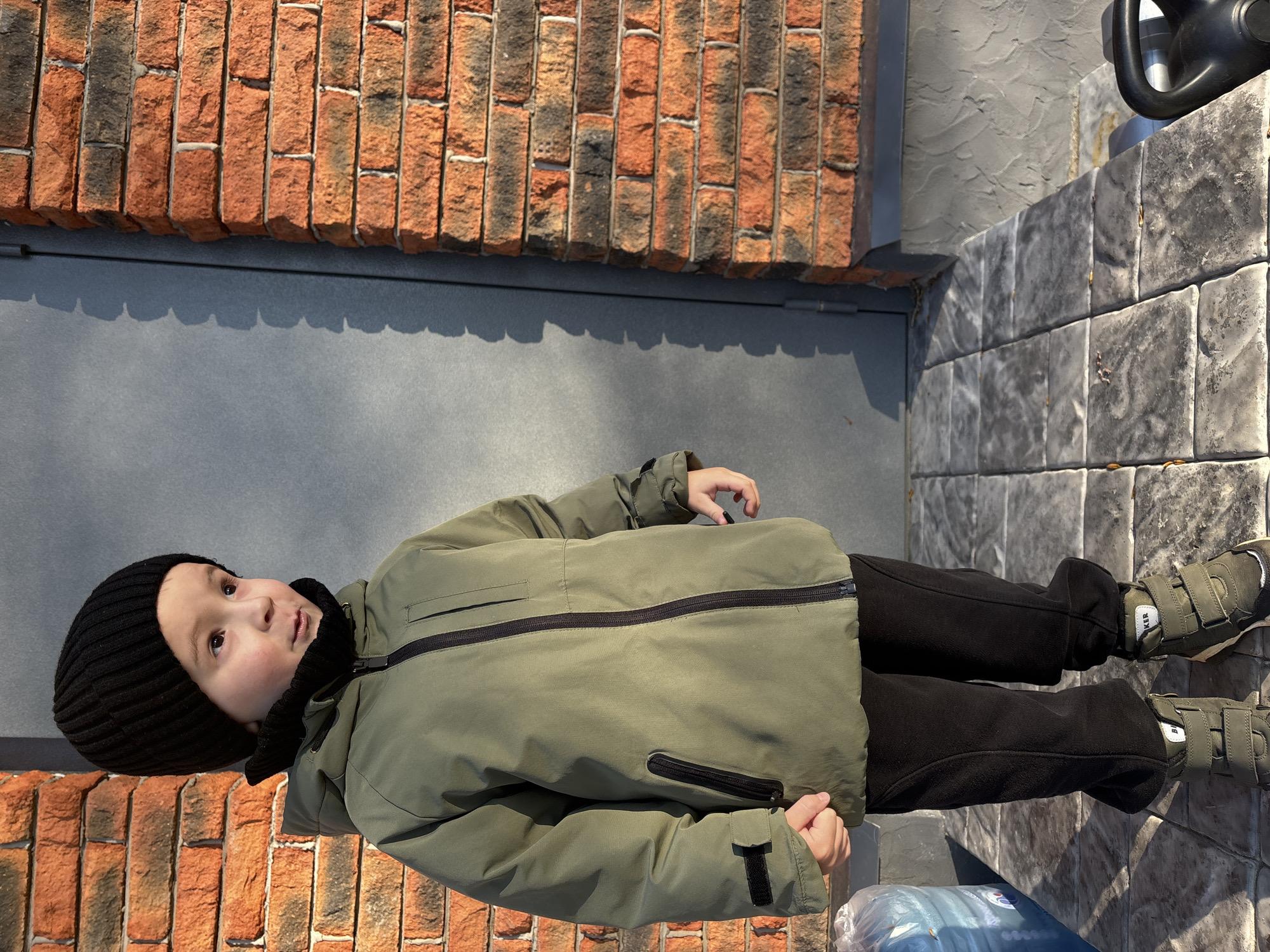 A young child, likely a boy, is depicted standing outdoors on a bright, sunny day in Chaikovsky, Russia. The child is dressed for cold weather, wearing a black knit beanie, an olive-green hooded winter jacket with black zippers, black pants, and olive-green shoes with three velcro straps. The word "BIKER" is visible on the velcro strap of the child's right shoe. The child looks upwards and slightly to the right with an open mouth, suggesting an expression of curiosity or engagement with something out of frame.
The background features a wall made of red and dark grey bricks on the left, which casts distinct scalloped shadows onto a dark grey wall or door immediately behind the child. The ground is covered with grey, stone-patterned paving slabs. On the right side of the image, partially visible on the paving, is a black watering can or similar garden tool. A translucent plastic bag or bundle is visible at the bottom left, near the child's feet. The strong shadows indicate direct sunlight, possibly in the morning or late afternoon. The overall scene captures a candid outdoor moment of a child.
