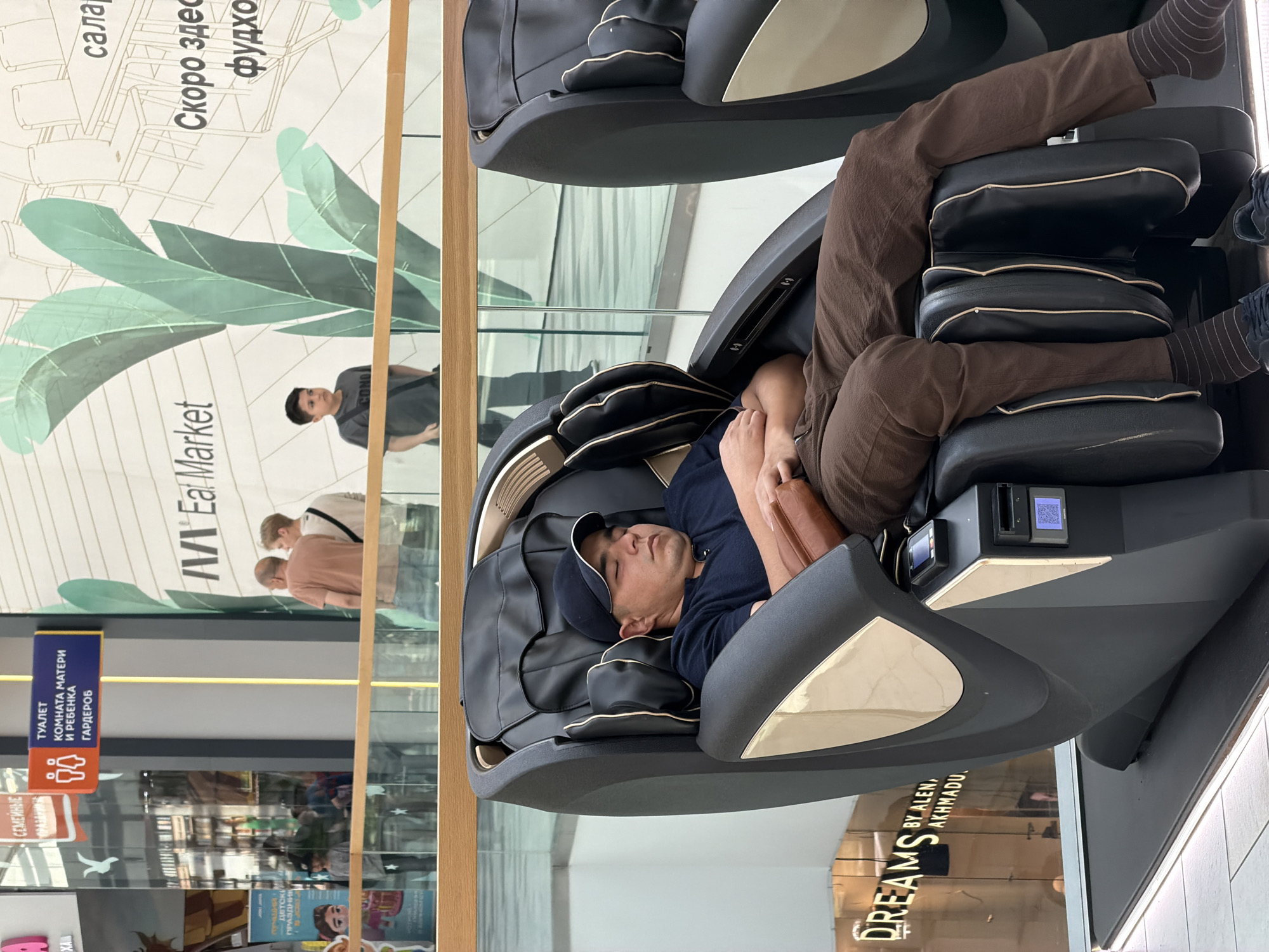 Here is a forensic description of the image:
**Scene:** The image appears to be taken inside a shopping center or mall in Kommunarka, Russia, as determined from Cyrillic script on signage visible in the background.
**Subject:** A person is relaxing, presumably asleep, in a high-end massage chair. They are wearing brown pants, socks, and a dark blue shirt. A baseball cap is on the head.
**Objects:** The massage chair is black with beige trim. It appears to be a sophisticated model, possibly pay-per-use, given the payment interface on the chair's armrest, which includes a screen and card reader. Background signage includes "M Eal Market," "Dreams by Alen, Akhmadu," and indicators for "Toilet", "Mother and Child Room", and "Wardrobe" facilities in Cyrillic. There are also advertisements for "Children's Holiday" visible.
**People:** Several figures are discernible through the glass, including two or three people standing near "M Eal Market". There's a person wearing a grey shirt.
**Environment:** The flooring is light-colored. The light indicates an indoor public space with heavy foot traffic.
**Actions/Interactions:** The main action is the individual sleeping in the massage chair. There is no direct interaction apparent between the person in the chair and the people visible in the background. The location and signage suggest a commercial environment designed for consumers.