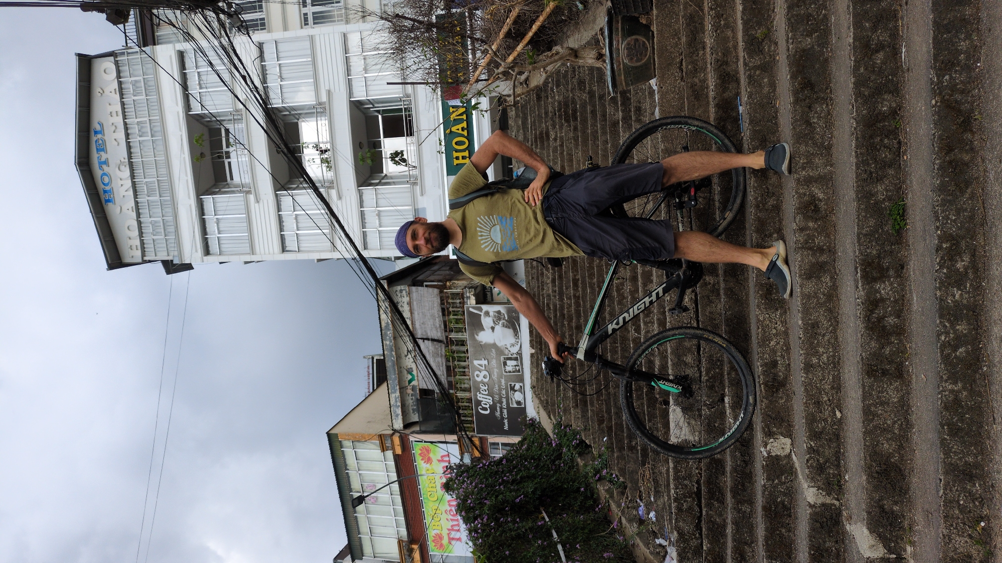 A man is posing with a mountain bike on concrete steps in Da Lat, Vietnam. The man is wearing a green t-shirt, dark shorts, and a blue head covering. He has one hand on his hip and the other on the handlebars of the bicycle. The bike is a dark color with a green accent. The man's legs stretch across the frame to rest on the steps behind him.
Behind him, a building reads "HOTEL HOAN." A sign for "Coffee 84" is partially visible, showing Vietnamese script and an image of a coffee cup. Another sign with "Bế Thiên" is slightly obscured by purple flowers in the lower left. Overhead, power lines crisscross against a cloudy sky. The steps are weathered, and the scene is tilted to the right.