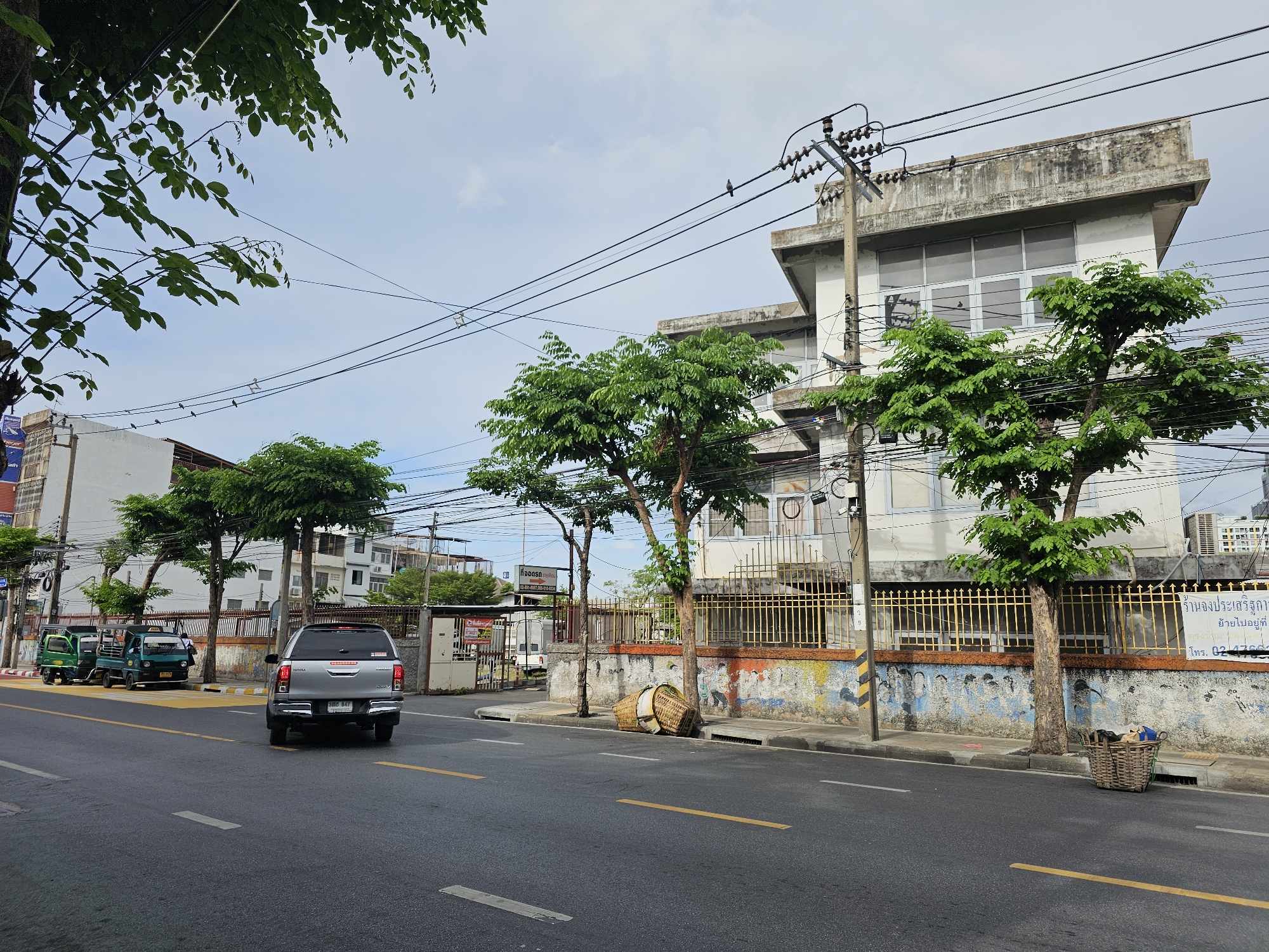 This is a street scene in Bangkok, Thailand, on a cloudy day. In the foreground, a gray pickup truck is driving away from the camera on a paved road with yellow and white lane markings. To the left of the road, there are three small green and yellow vehicles parked. Beyond these vehicles and the road, there is a sidewalk with a low, colorful wall adorned with graffiti. Behind the wall, there are several trees with green foliage. A tall building with several windows and a concrete roof is visible behind the trees and wall, with electrical wires and poles in front of it. Two woven baskets are placed on the ground near the sidewalk, one containing some items. The sky is partly cloudy.
