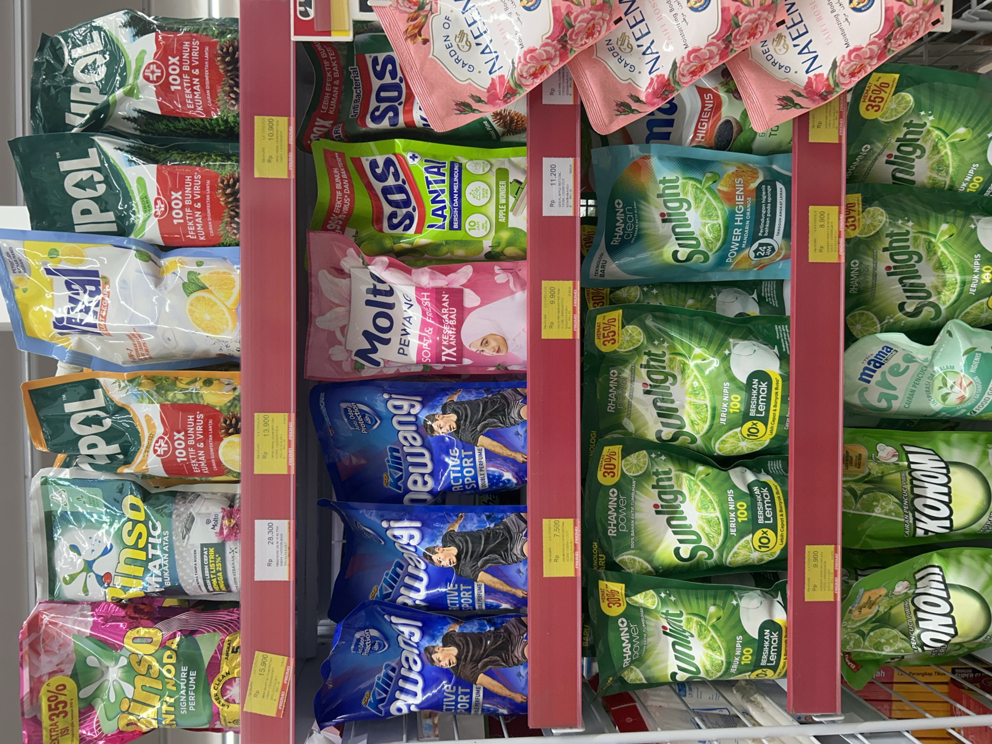 This media file captures a well-stocked aisle in what appears to be a supermarket or retail store in Payadua, Indonesia, displaying various cleaning and laundry products. The items are neatly arranged on red shelves, primarily in flexible pouch or sachet packaging, indicating refill or single-use formats.
The subjects are diverse household products, including floor cleaners, dishwashing liquids, laundry detergents, and fabric softeners. Prominent brands like "Vipol," "Rinso," "Molto," "SOS," "Sunlight," "Ekonomi," "Mama Lime," and "Klin Pewangi" are visible. Some products, like "Na'een," appear to be body care items.
Notable details include numerous yellow price tags displaying prices in Indonesian Rupiah (Rp), such as Rp 15.900, Rp 28.300, Rp 10.900, Rp 9.900, and Rp 8.900. Many tags feature "PROMO" labels and indicate discounts like "HEMAT 30%" or "HEMAT 35%," with some offering "EXTRA 35% ISI" (extra 35% content).
Visible text highlights product features and scents:
- "Vipol" boasts "100X EFEKTIF BUNUH KUMAN & VIRUS" (100X effective kill germs & viruses) for "CAIRAN DISINFEKTAN LANTAI" (floor disinfectant liquid), with pine or lemon variants.
- "Sunlight" dish soaps emphasize "JERUK NIPIS 100" (lime 100) and "10X BERSIHKAN LEMAK" (10x clean grease), with variants like "POWER HIGIENIS MANDARIN ORANGE" and "RHAMNO POWER 100% BAHAN AKTIF TUMBUHAN" (100% plant-based active ingredients).
- "Molto" fabric softener is advertised as "PEWANGI SOFT & FRESH 7X KESEGARAN ANTI BAU" (soft & fresh fragrance, 7x freshness anti-odor).
- "SOS Lantai" floor cleaner comes in "APPLE WONDER" scent.
- "Rinso" offers "ANTI NODA SIGNATURE PERFUME" (stain remover signature perfume) and "MATIC BUKAAN ATAS" (top-load automatic).
- "Klin Pewangi" is available in "ACTIVE SPORT DOUBLE PERFUME" with "Anti Odor Protection 48h."
- "Na'een" specifies "TAIFI ROSE MOISTURIZING BODY."
No people are visible in the scene, and the setting appears to be indoors with artificial lighting. The overall impression is one of a busy retail environment offering various household essentials with ongoing promotions.