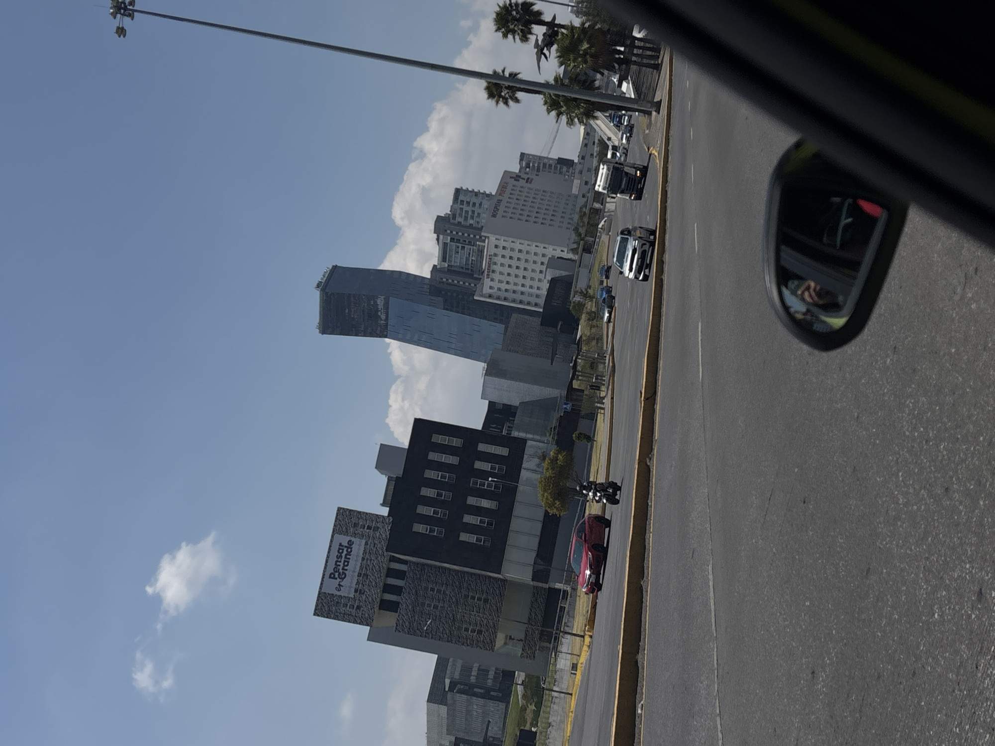 A sideways view from a car shows a street in Puebla, Mexico, with a cityscape along the left. Prominent buildings include a glass skyscraper and a darker, block-like building with "Pensar en Grande" on its facade. A highway runs to the right, with cars and a motorcycle in the lanes. Palm trees line the road further down. The sky is blue with scattered clouds. The car's side mirror is visible in the upper right corner of the frame.