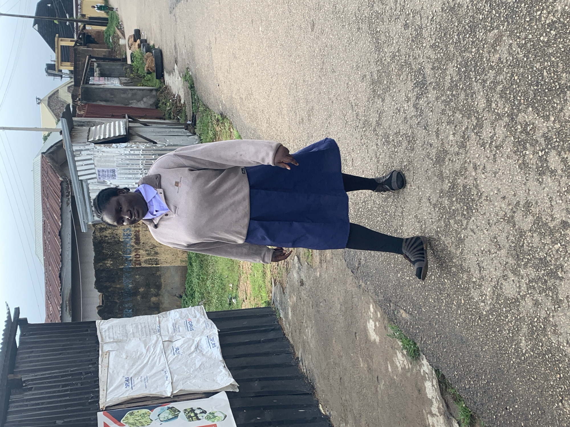 A young Black woman, appearing to be a student, walks along a paved road on a cloudy day. She is wearing a light brown long-sleeved shirt over a light blue collared shirt, a dark blue knee-length skirt, black leggings, and dark grey strapped sandals. Her hair is pulled back in braids. The road is lined with corrugated metal structures and residential buildings with various roofing materials, including corrugated metal and asphalt shingles. Some of the buildings appear weathered and in disrepair. Piles of tires are visible along the roadside, and overgrown vegetation is present in patches. The woman is walking towards the right of the frame, looking forward. In the background, another individual, possibly a student in uniform, is visible further down the road. The scene suggests a modest urban or semi-urban environment in Jos, Nigeria, characterized by its architecture and streetscape.