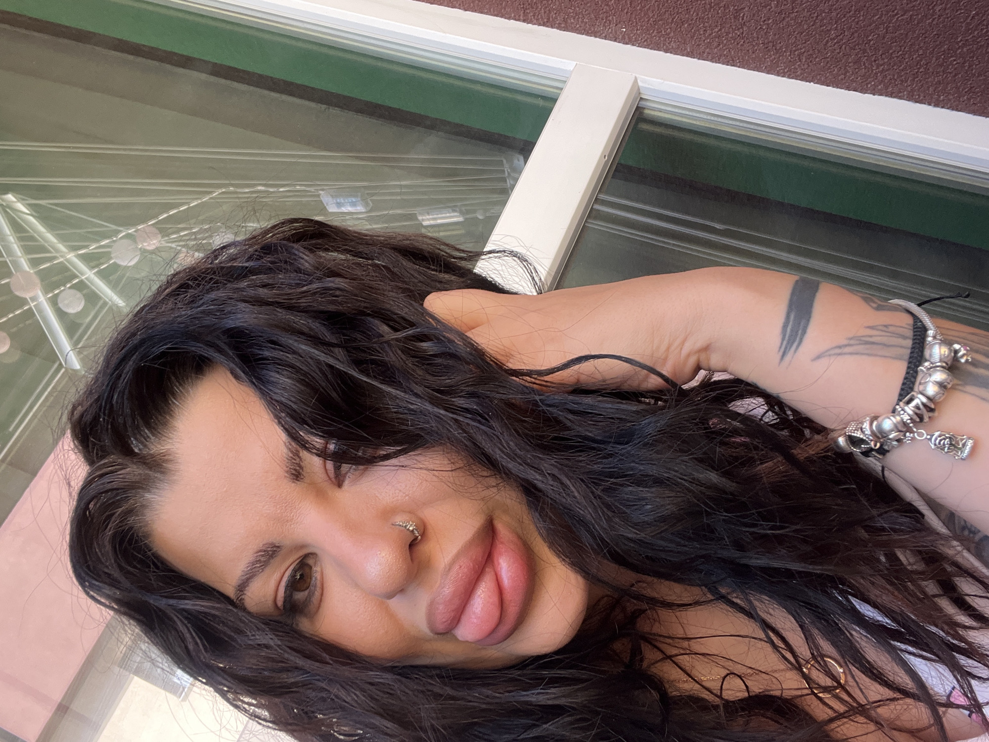 A close-up, tilted selfie shows a woman with long, dark wavy hair. She has fair skin and facial features including brown eyes, a nose ring, and full lips. Her head is positioned towards the lower left corner of the frame. Her right hand is raised and resting behind her head, displaying a tattoo on her forearm and multiple bracelets on her wrist. The background consists of a window with a white frame. Light reflecting off the window suggests an outdoor environment with varying colors. The location within the city of Plovdiv, Bulgaria, cannot be determined from the visual information provided.