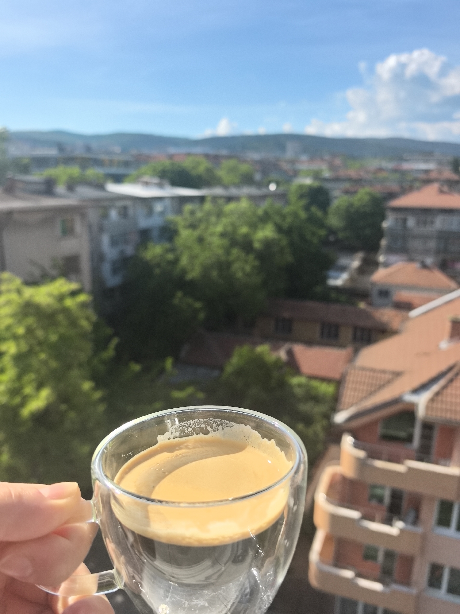 A hand holds a clear glass cup of coffee, with visible crema, in the foreground. The background showcases a cityscape under a bright blue sky, punctuated by trees and buildings. The architecture includes tiled roofs and multi-story buildings. In the distance, a mountain range is visible. The location is Stara Zagora, Bulgaria, discernible from the common architectural characteristics.