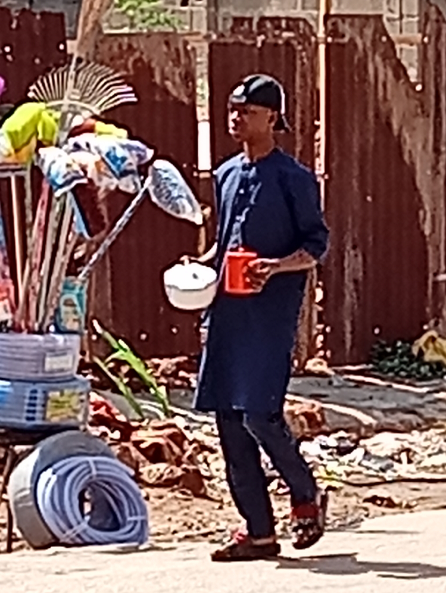In Maiburiji, Nigeria, a person wearing a dark blue tunic and dark pants, with a black baseball cap tilted backward on their head, is walking on a light-colored paved surface. They are holding a small white bowl in their left hand and a red mug in their right hand. Behind them, there's a chaotic scene: a metal stand supporting various items, including rakes, fabrics, and containers, as well as stacked blue plastic baskets and coiled blue tubing on the ground. A weathered corrugated metal fence forms the backdrop. Debris is scattered on the ground.