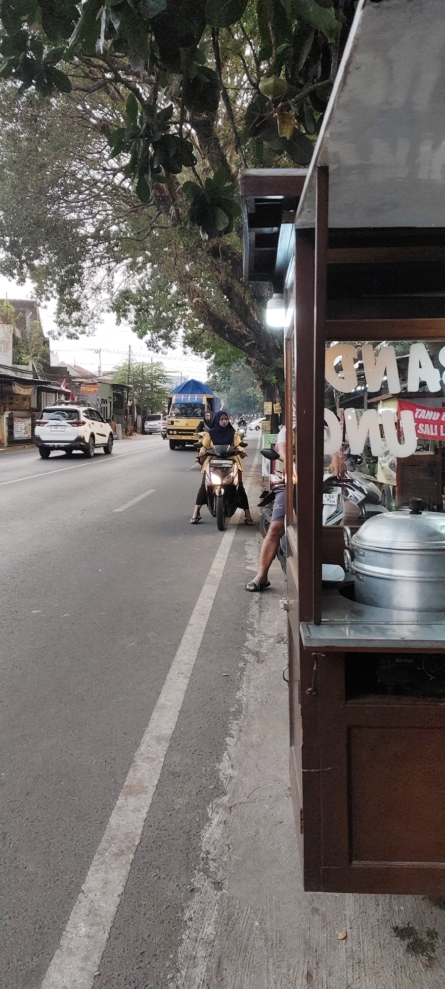 A street-level, eye-level shot captures a bustling street scene in Malang, Indonesia. The foreground is dominated by a dark brown food stall with a light-colored, angled awning. A large, silver steaming pot sits prominently on a shelf within the stall. Through the transparent front of the stall, white lettering on glass is visible, though partially obscured and difficult to decipher.
The street itself is paved with asphalt and marked by a white broken line. On the road, a woman wearing a yellow hijab and a dark outfit rides a scooter. Her feet are bare and clad in flip-flops. Further down the street, a white SUV is driving away from the viewer, and a yellow truck with a blue tarp on its back is also present.
The background reveals a line of buildings and lush trees lining the street, with their branches extending over the road. The sky appears overcast, suggesting it might be morning or late afternoon. The overall atmosphere is one of everyday urban life in Malang, with traffic and commercial activity in full swing.