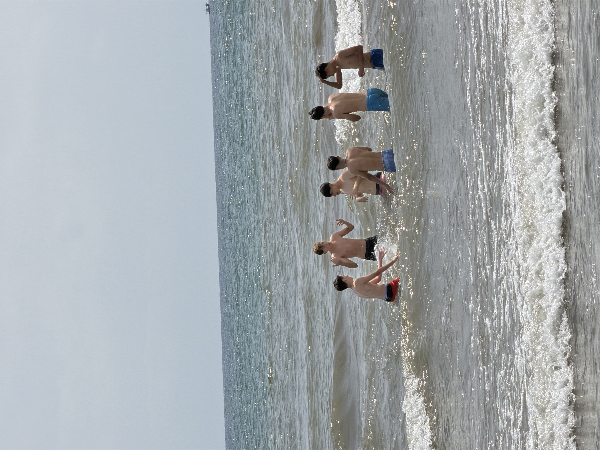 The image shows six young males in the ocean at waist-deep water. They are all shirtless and wearing swim trunks. The ocean is calm with small waves and the sky is clear and overcast. The setting appears to be a beach on a sunny day.